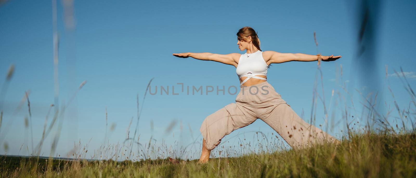 Young Woman Practicing Yoga Outdoors in the Field with Blue Sky on the Background by Romvy