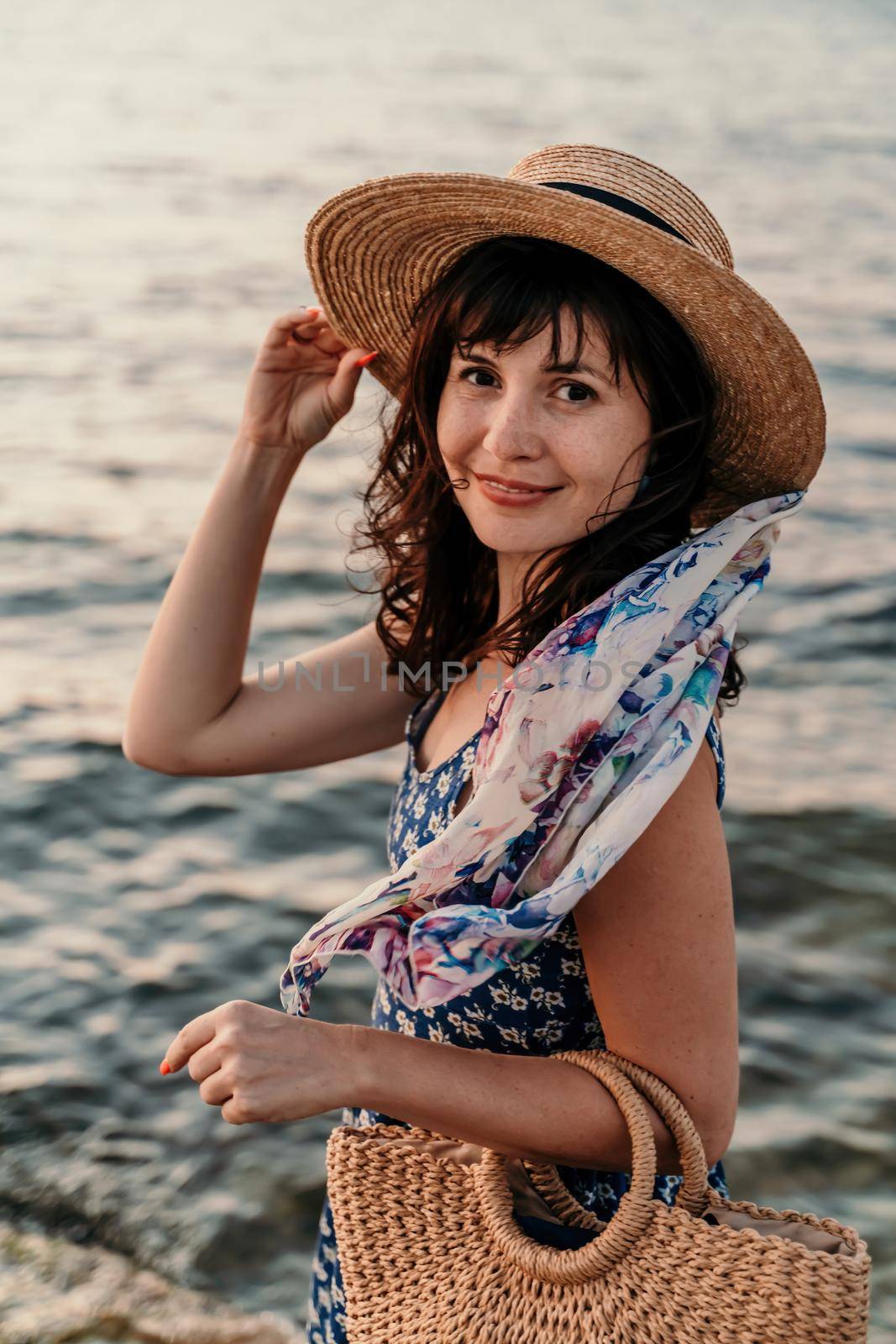 A woman in a dress, hat and with a straw bag is standing on the beach enjoying the sea. Happy summer holidays.