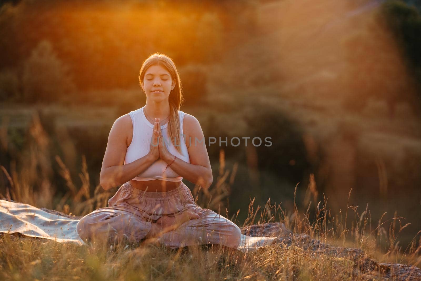 Happy Young Woman with Folded Hands and Closed Eyes Sitting on the Mat and Meditating Outdoors at Sunset by Romvy