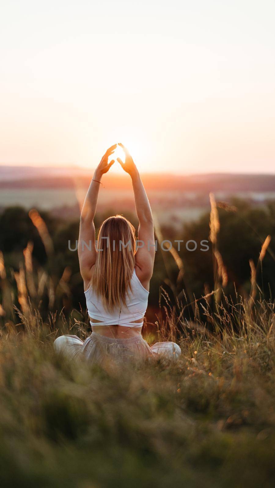 Back View on Woman Sitting in Meditation Yoga Pose and Catching Sun by Her Hands at Sunset Outdoors