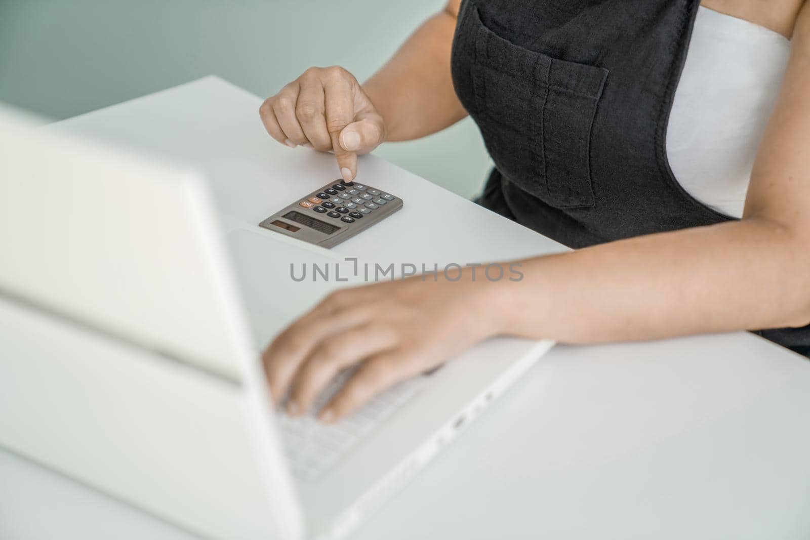 Young woman is sitting at her laptop and doing calculations on calculator. Hands, calculator, and laptop close-up. Concept of financial and project calculations and accounting.