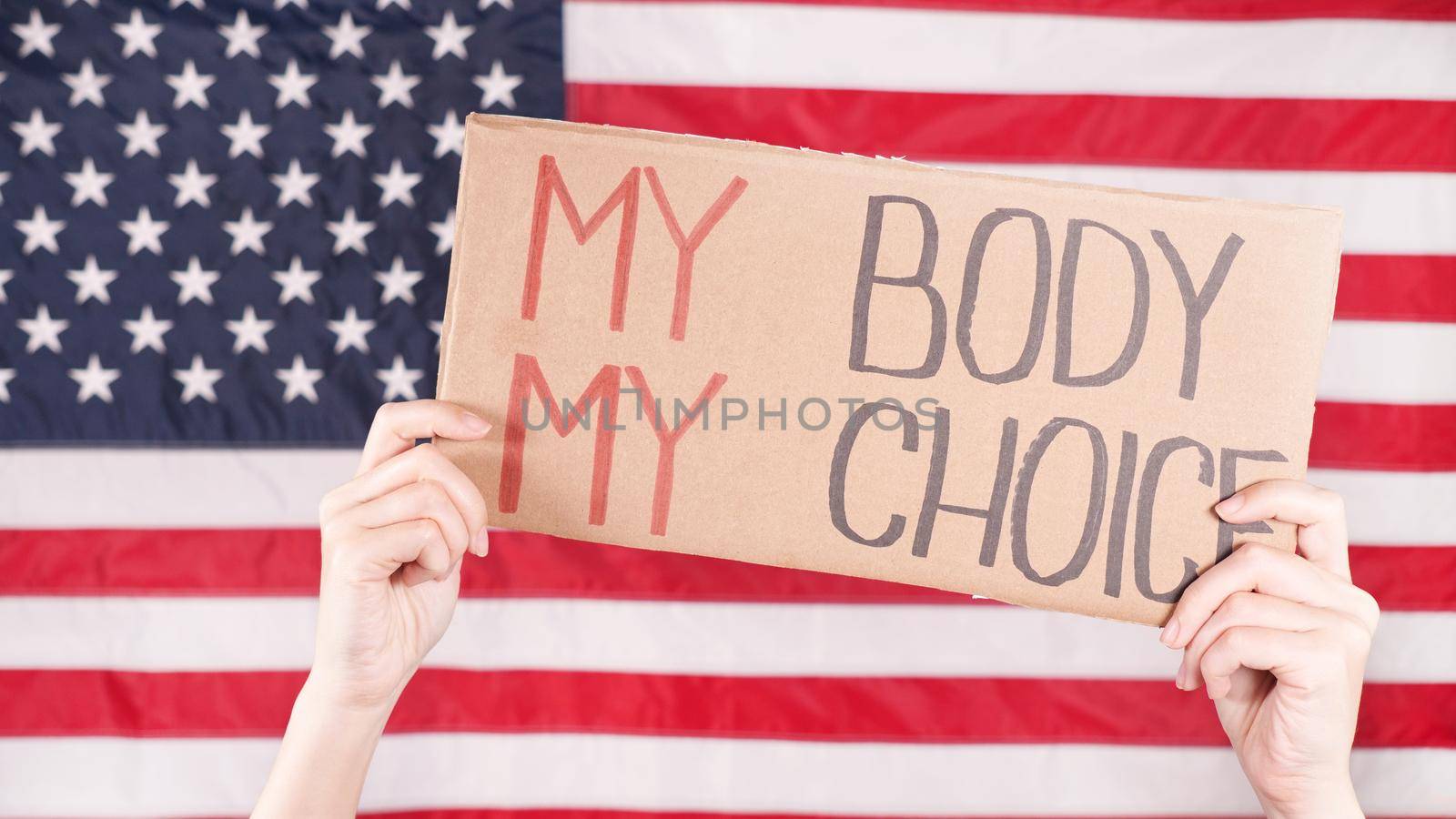 Young woman protester holds cardboard with My Body My Choice sign against USA flag on background. Girl protesting against anti-abortion laws. Feminist power. Equal opportunity Womens rights reedom. by JuliaDorian
