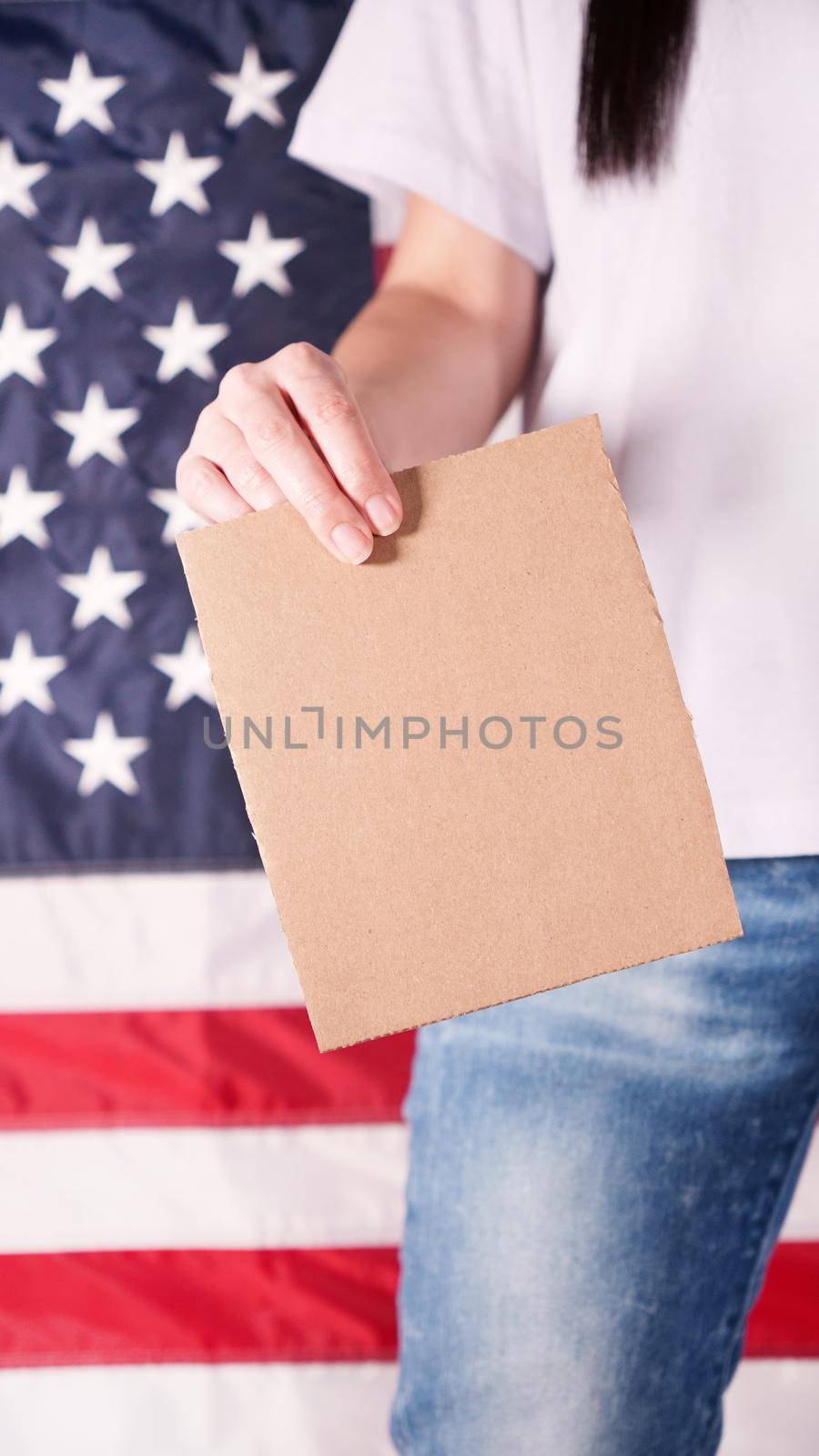 Young woman holds empty cardboard with Space for Text sign against American flag on background. Girl protesting anti-abortion laws. Feminist power. Womens rights freedom. by JuliaDorian
