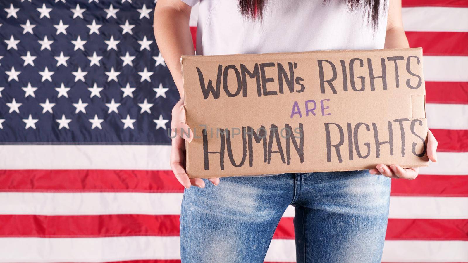 Young woman protester holds cardboard with Womens Rights Are Human Rights sign against USA flag on background. Feminist power. Equal opportunity Womens rights freedom. by JuliaDorian