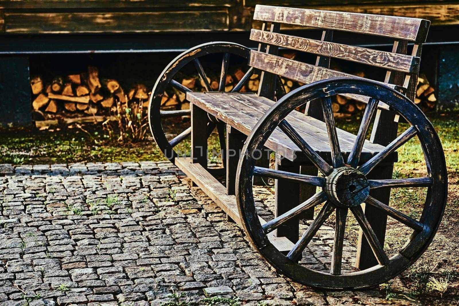 a long seat for several people, typically made of wood or stone. Wooden vintage bench with cart wheels on paving stones.