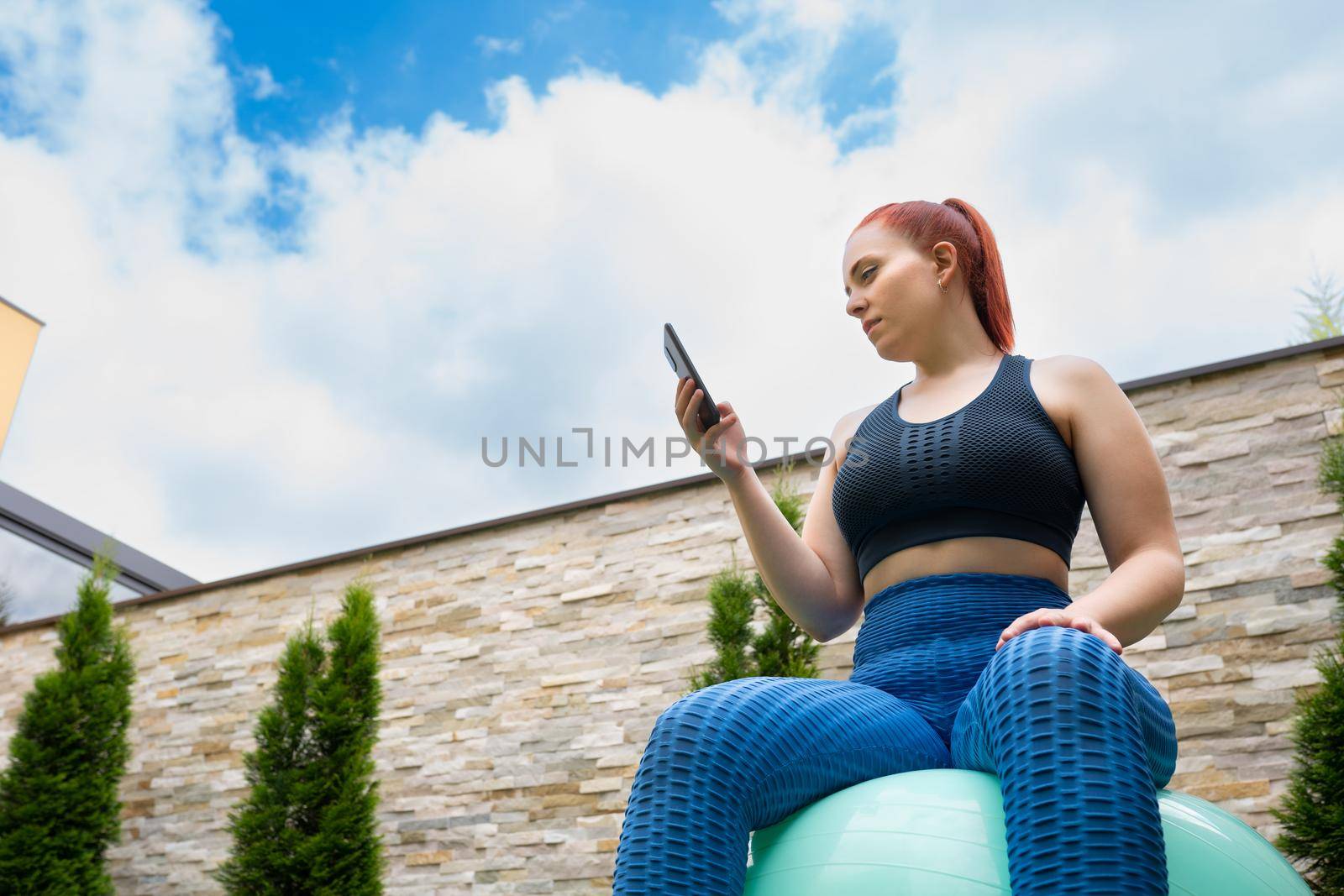 Attractive young fitness girl looking at her smart phone sitting on a pilates ball outdoors. girl exercising while sharing it on her social networks. health and wellness concept. by CatPhotography