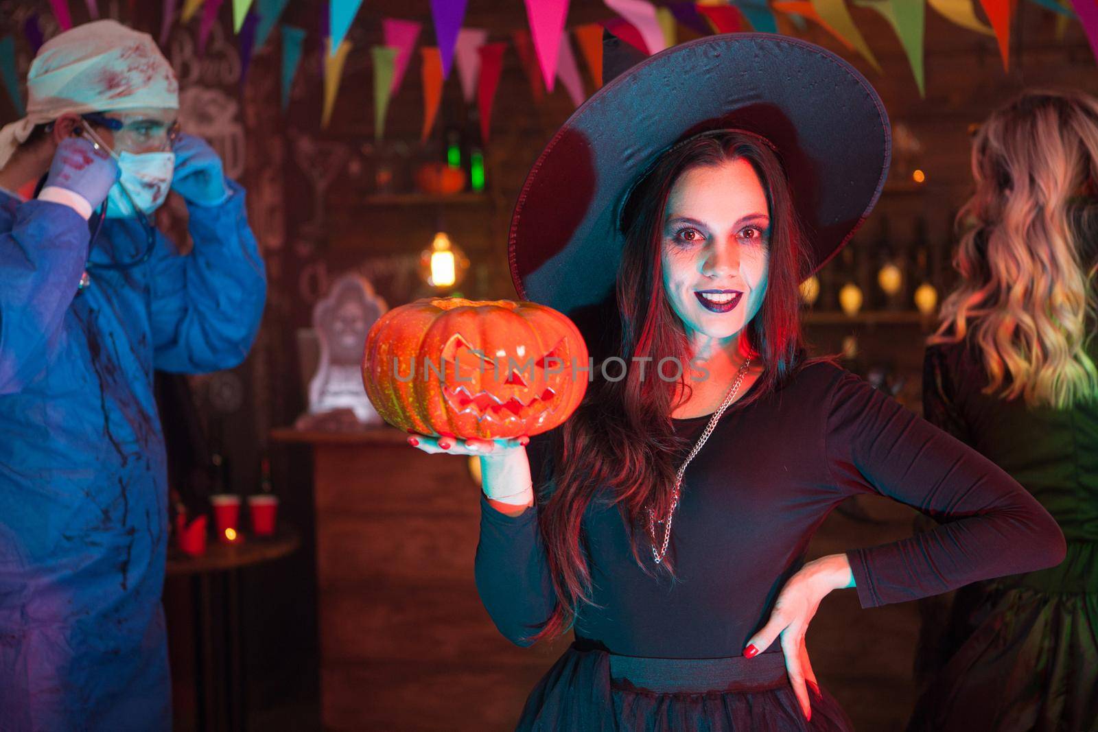 Attractive young woman dressed up like a witch for halloween posing in front of the camera. Creepy doctor in the background.