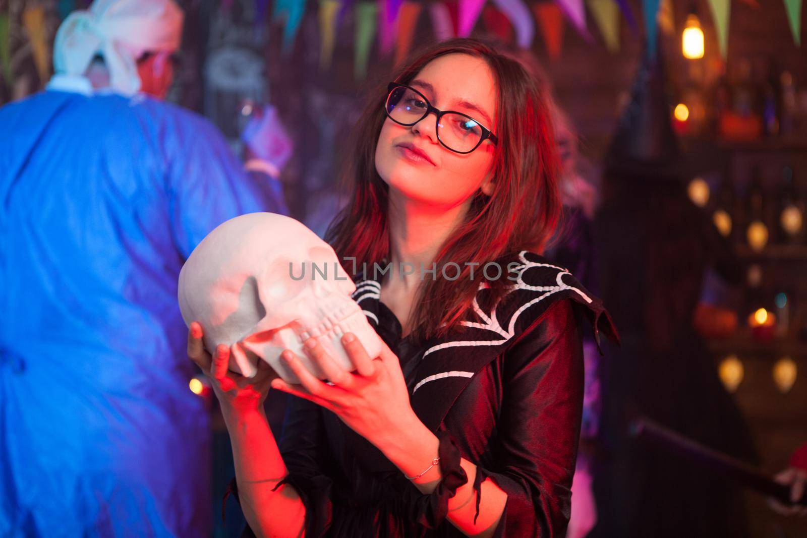 Pretty little girl dressed up like a witch holding a human skull at halloween party. Creepy man dressed up like a doctor in the background.