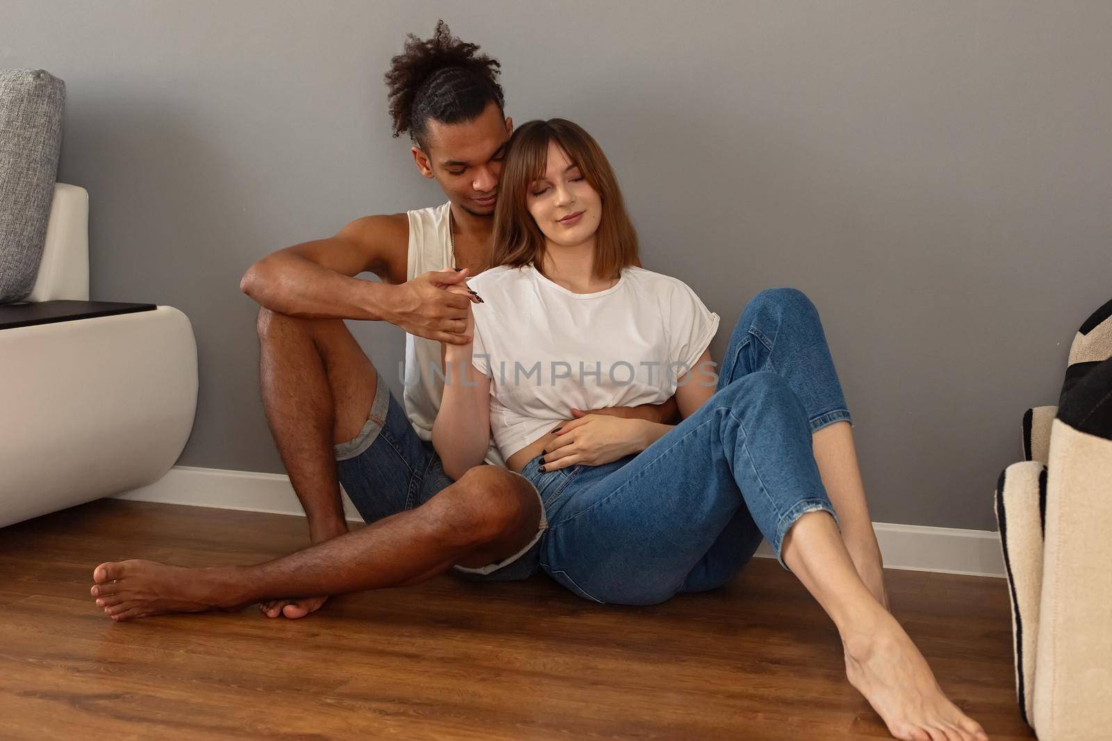 A multiracial gentle couple, a guy and a girl in white t-shirts and blue jeans, sit on the floor at home against a gray wall, hugging. Copy space