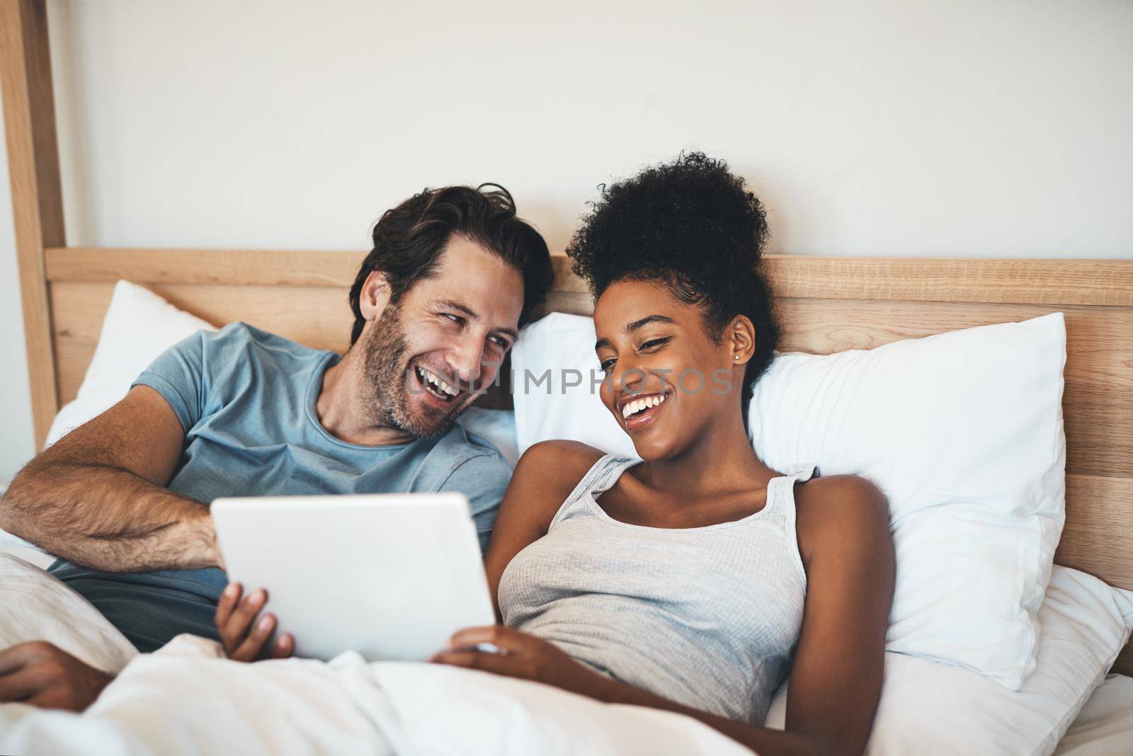 Happy, relaxed and carefree couple reading social media news on digital tablet and laughing in bed. Interracial husband and wife waking up together and browsing internet, sharing a funny online joke by YuriArcurs