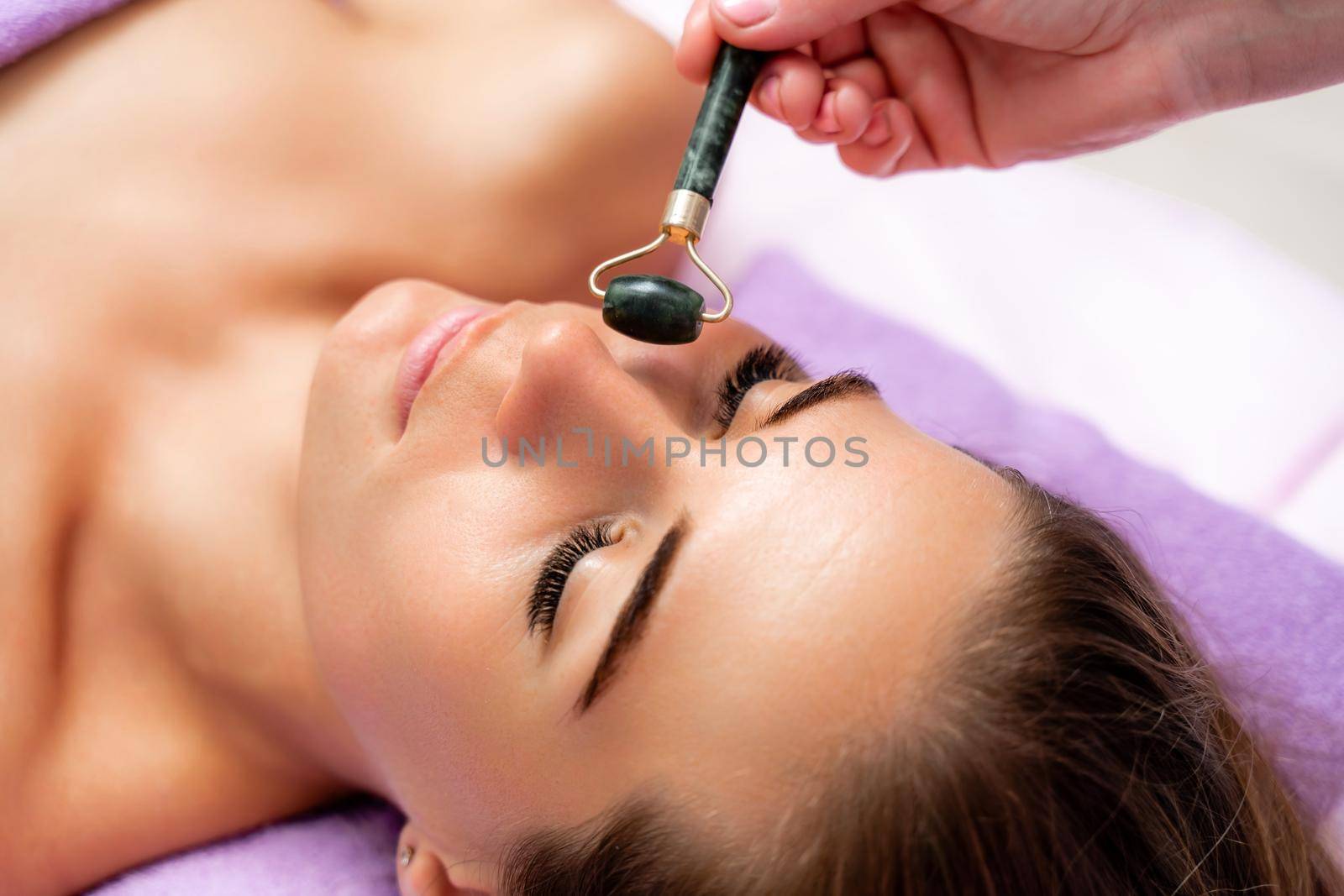 Relaxing massage. Side view european woman getting massage with jade face roller gouache in spa salon.