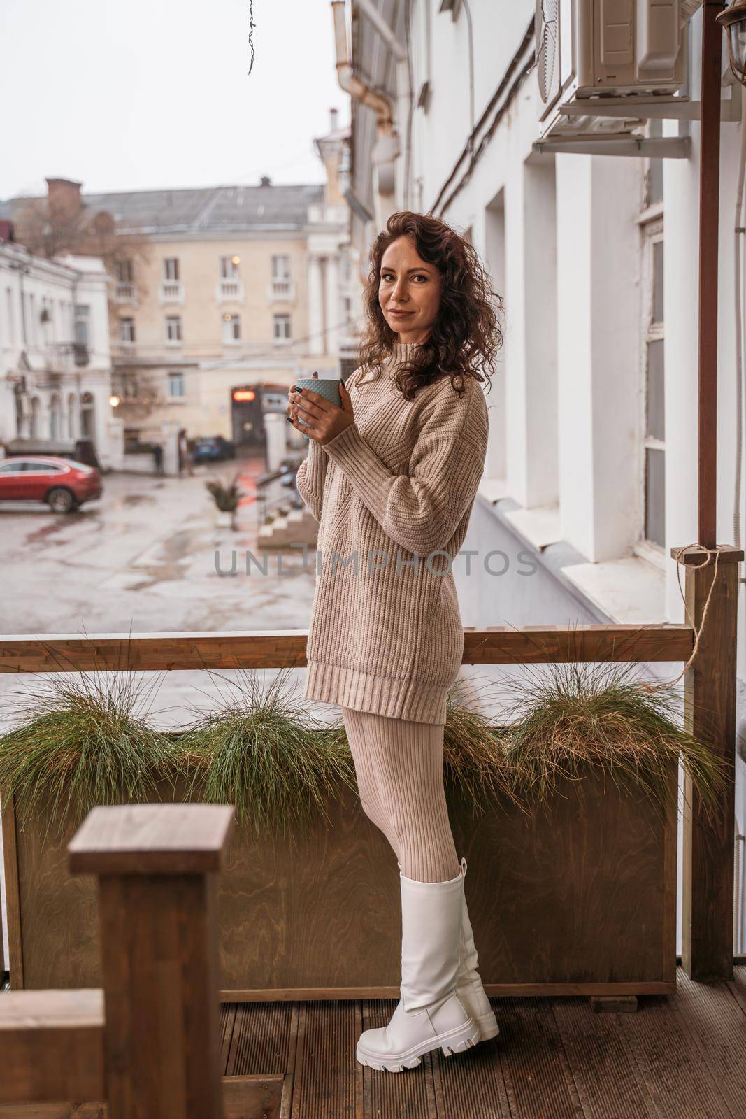 A middle-aged woman in a beige sweater with a blue mug in her hands is in a street cafe on the veranda by Matiunina