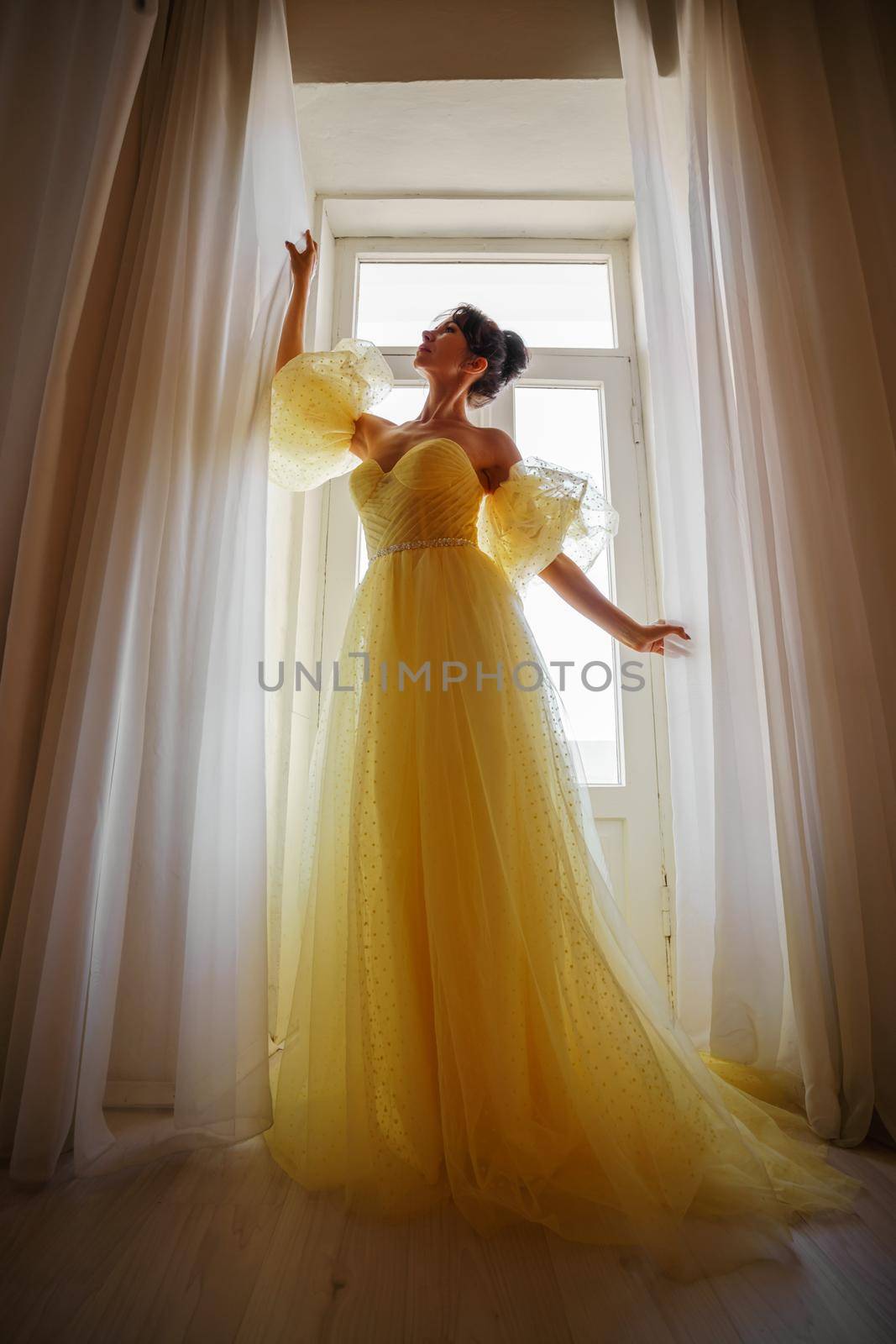 A woman's silhouette in a golden luxurious dress against the background of a window holds a curtain with her hands. Elegant lady in a yellow long silk dress with bare back, back view. by Matiunina