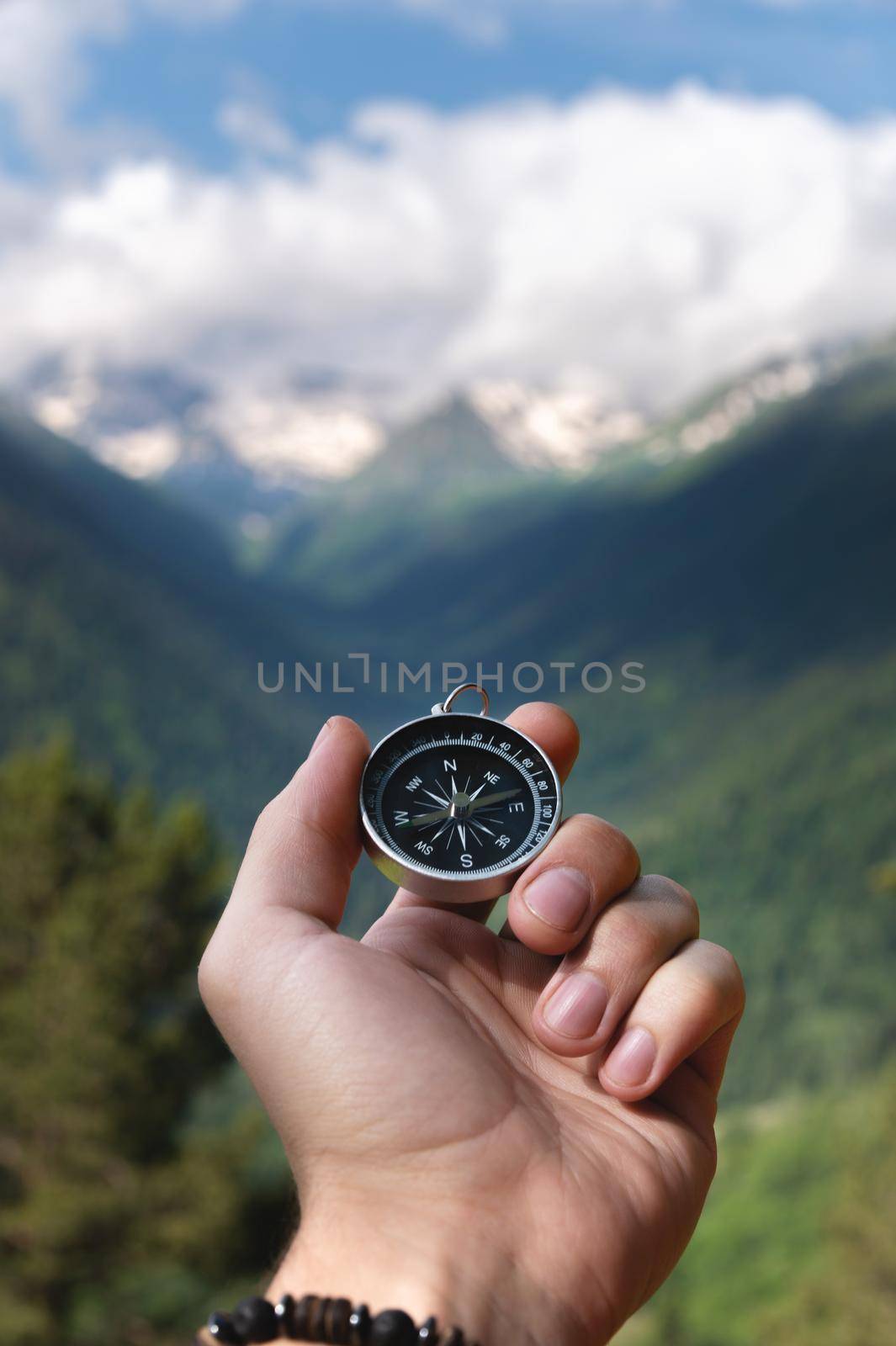 A hand with a compass against the backdrop of epic snow-capped mountains with clouds and a forest at the foot, close-up.