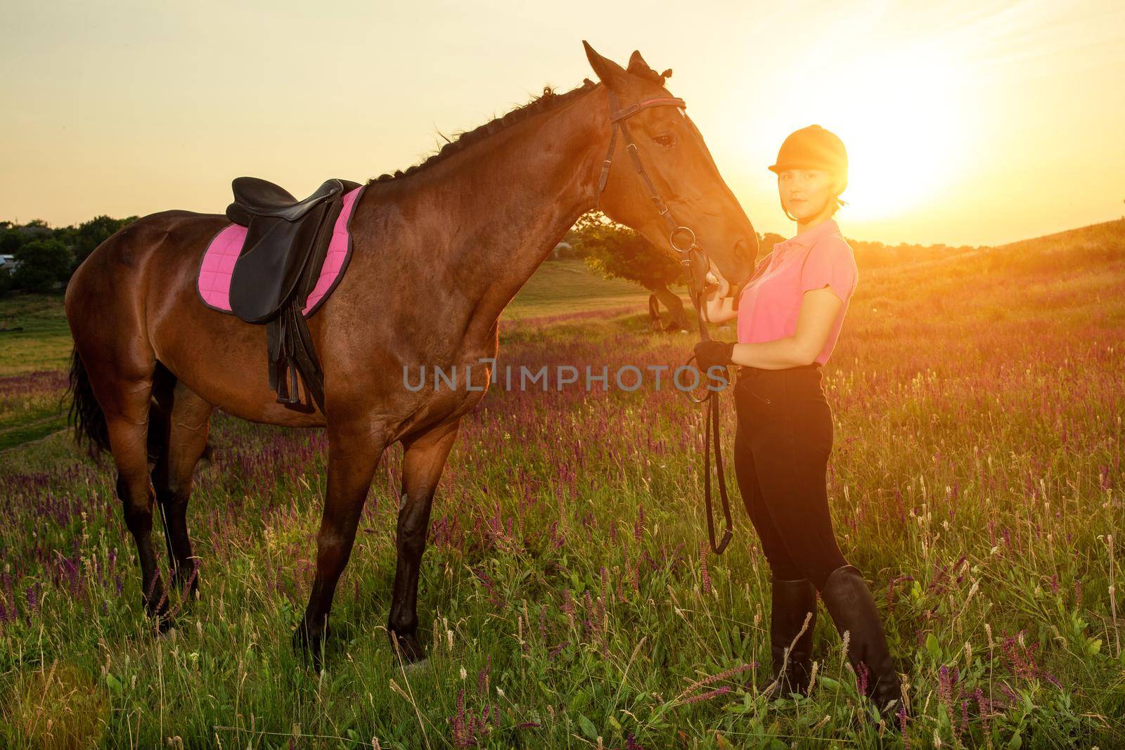 Beautiful smiling girl jockey stand next to her brown horse wearing special uniform on a sky and green field background on a sunset. Equitation sport competition and activity.