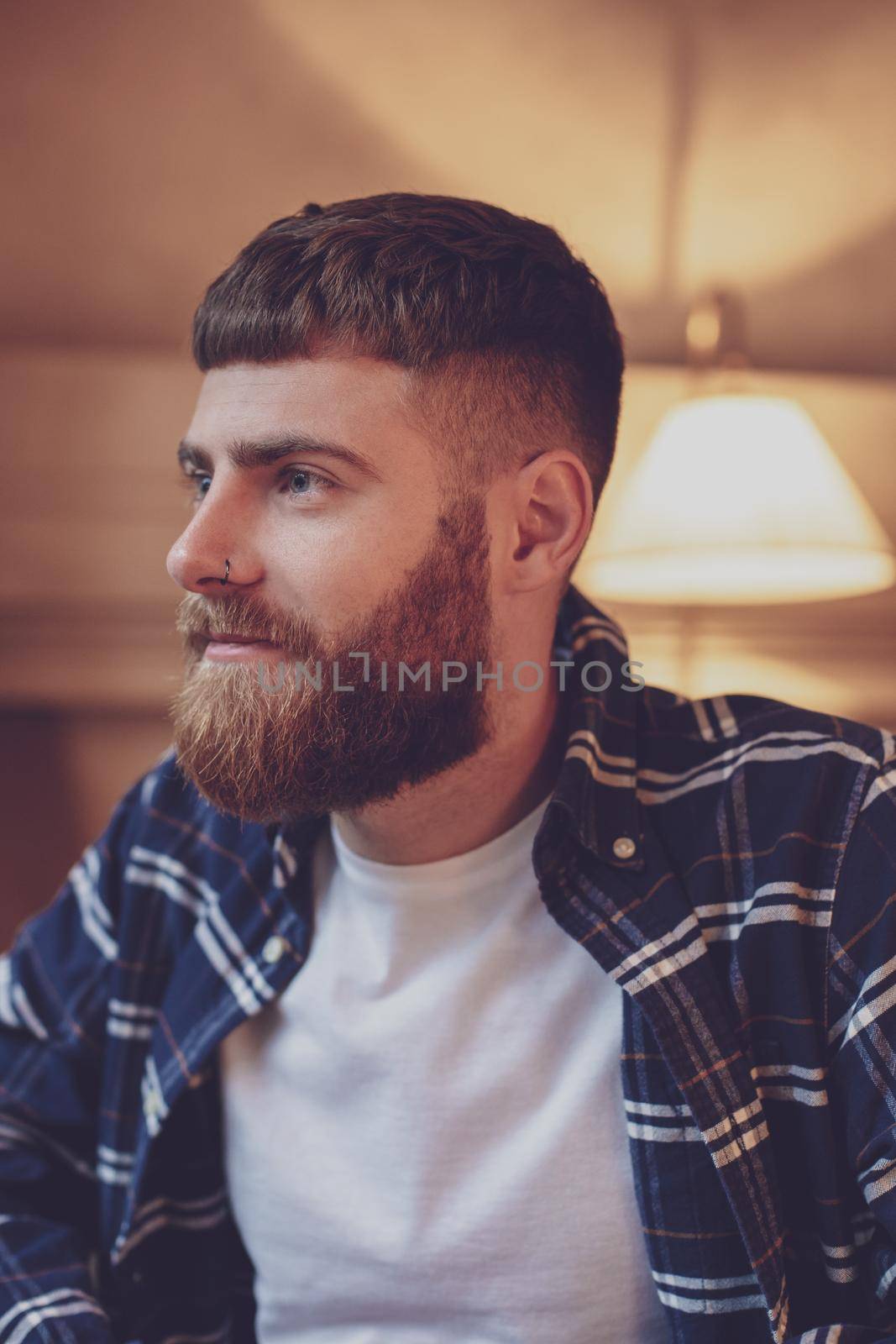 Portrait handsome bearded man wearing plaid shirt at modern cafe. Guy sitting in wooden chair and relaxing.