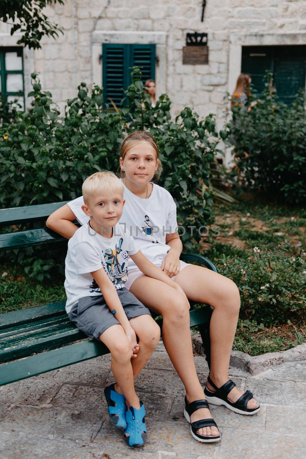A little boy with long hair and a girl are sitting on a park bench. Children talk while sitting in a pack on a bench. Boy and girl spend their free time outdoors. Portrait of a girl and a boy.