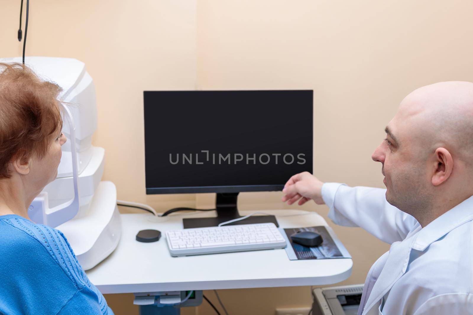 A male ophthalmologist explains the results of an eye examination to an adult woman, showing a drawing on a computer.