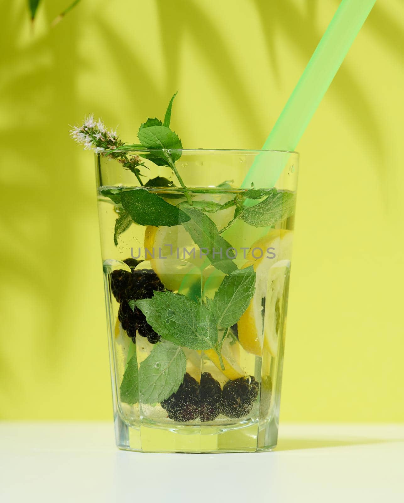 Transparent glass with lemonade, mint leaves, lemon slices and blackberries in the middle. Green background