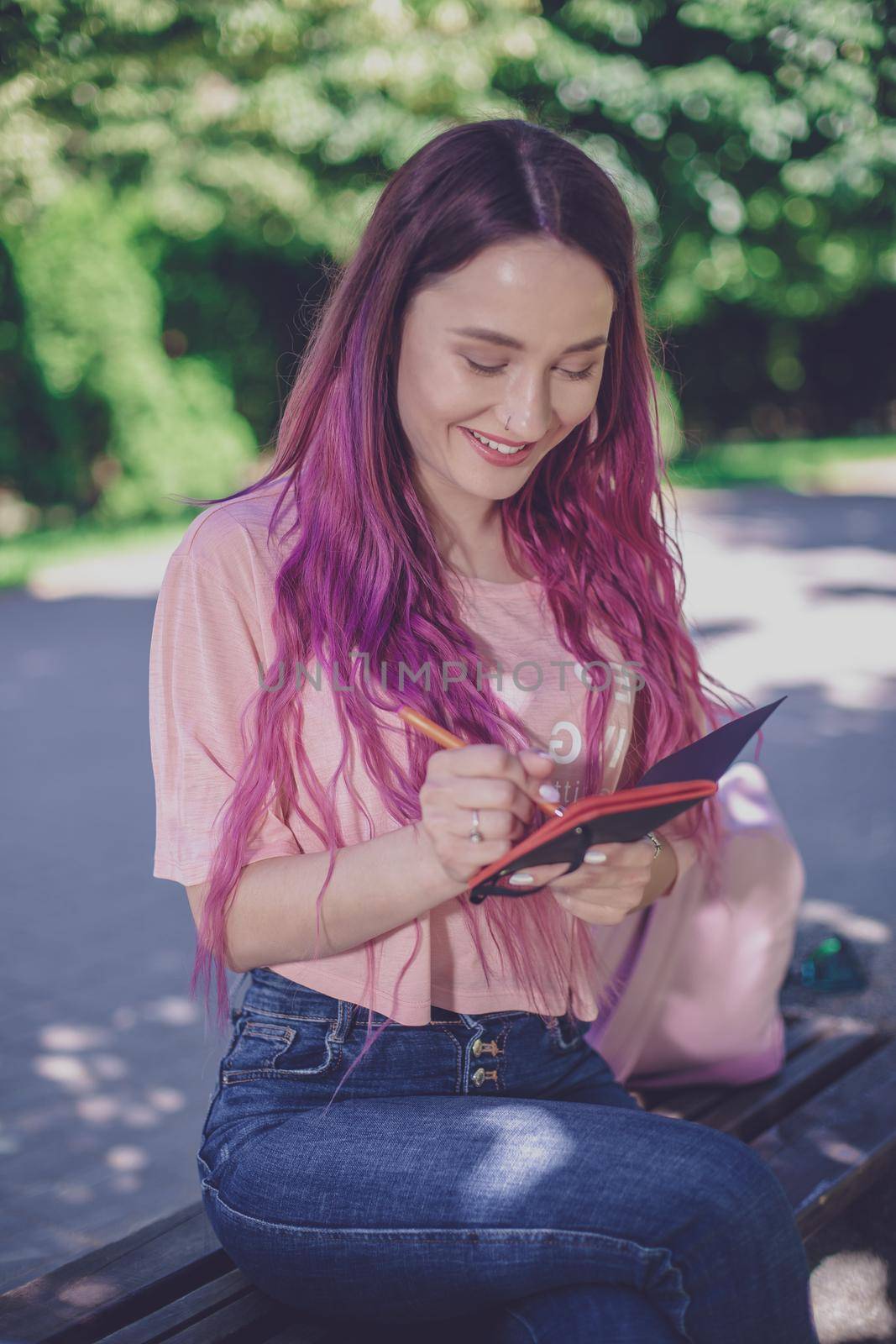 Woman writing in a notebook sitting on a wooden bench in the park. Girl working outdoors on portable computer, copy space. Technology, communication, freelance and remote working concept.