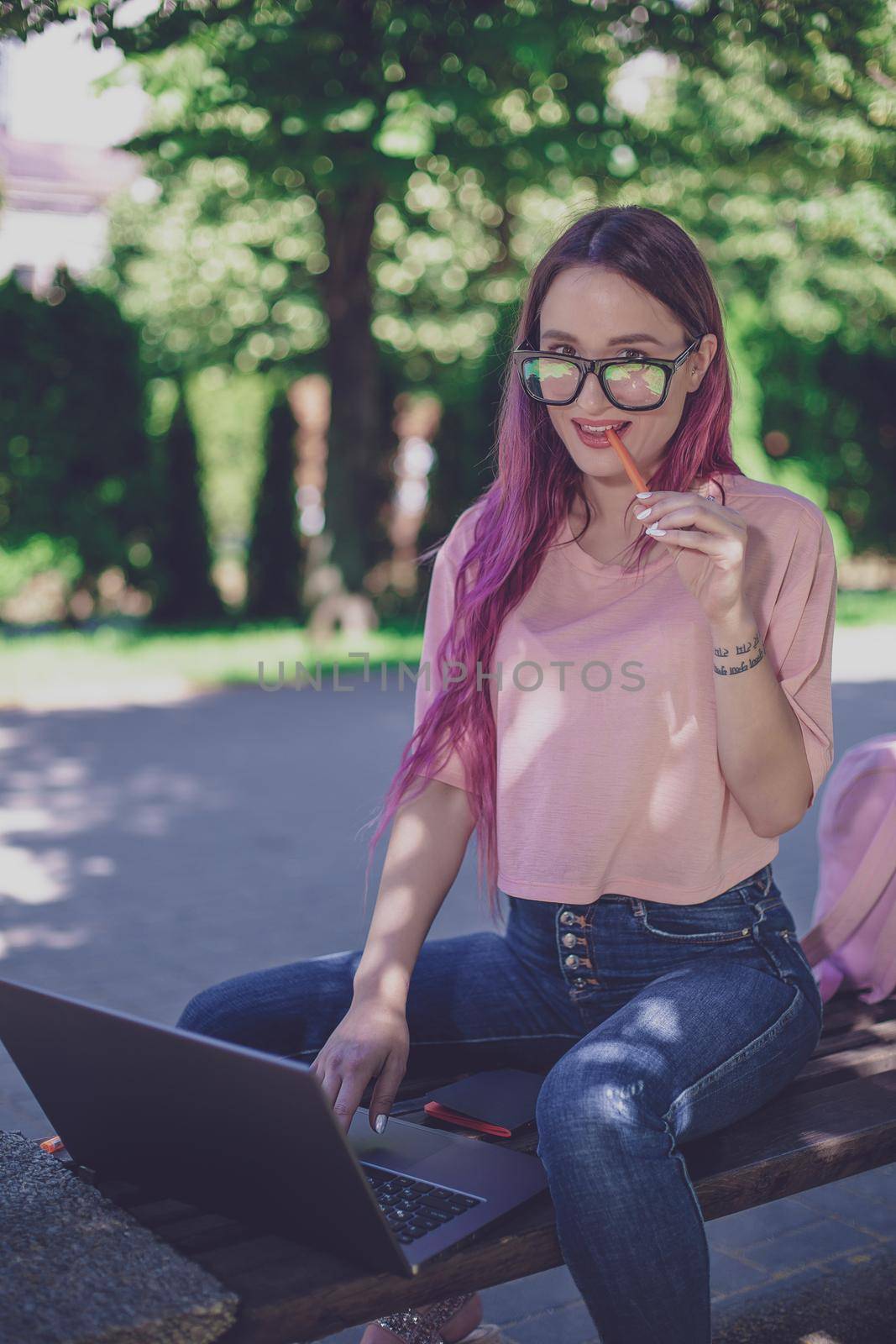 Young girl with pink hair is studying in the spring park, sitting on the wooden bench and browsing on her laptop