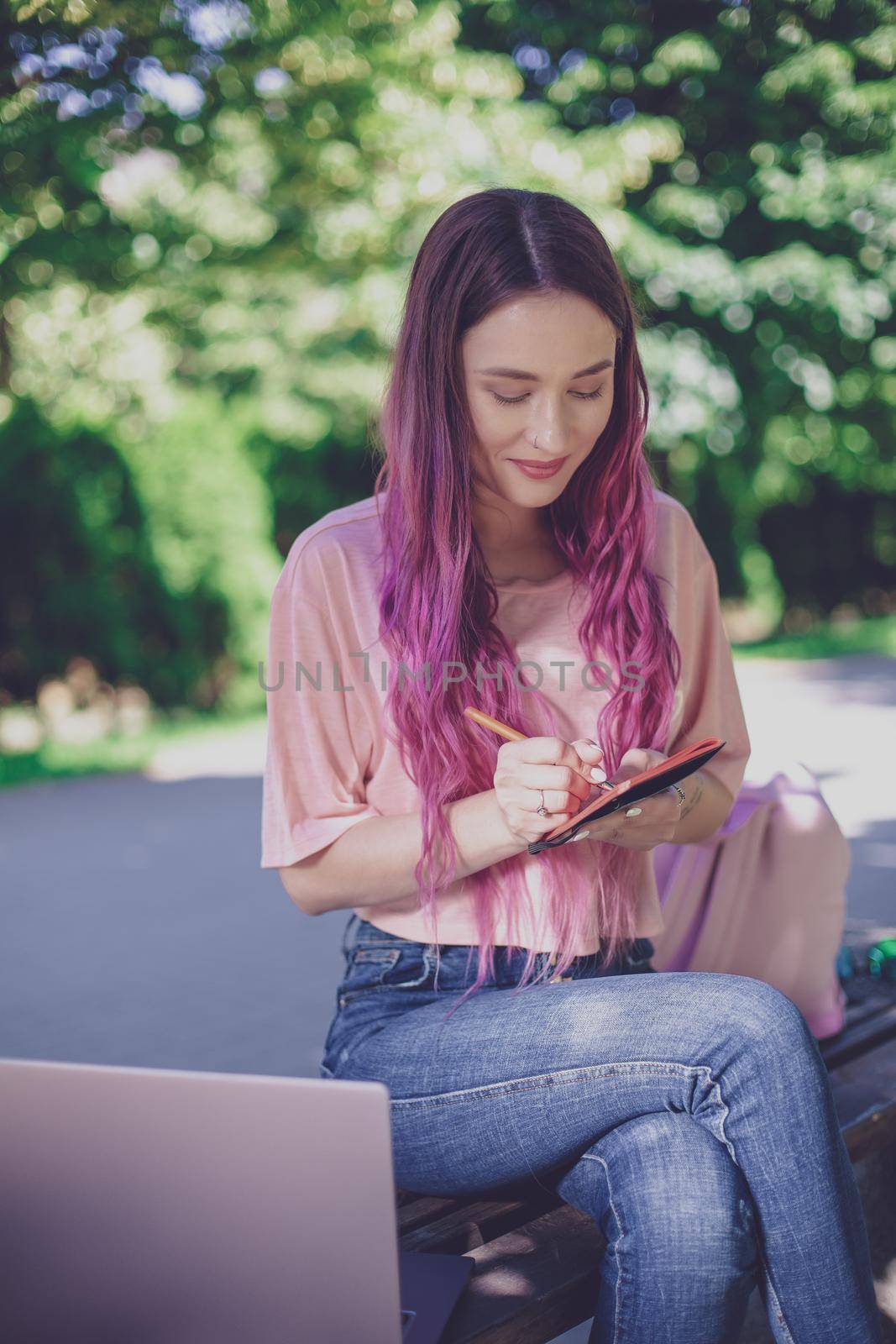 Woman writing in a notebook sitting on a wooden bench in the park. Girl working outdoors on portable computer, copy space. Technology, communication, freelance and remote working concept.