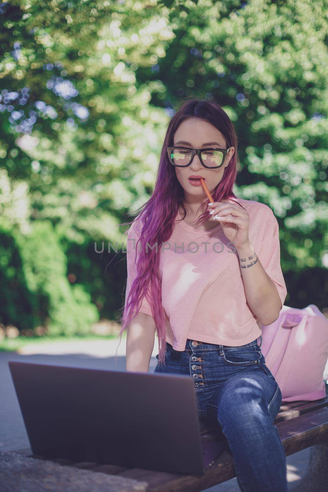 Young girl with pink hair is studying in the spring park, sitting on the wooden bench and browsing on her laptop