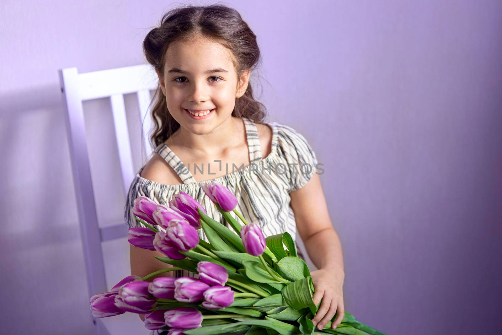 Beautiful little girl with a bouquet of tulips, sits on a white chair, against a purple wall, smiles, looks at the camera. Copy space