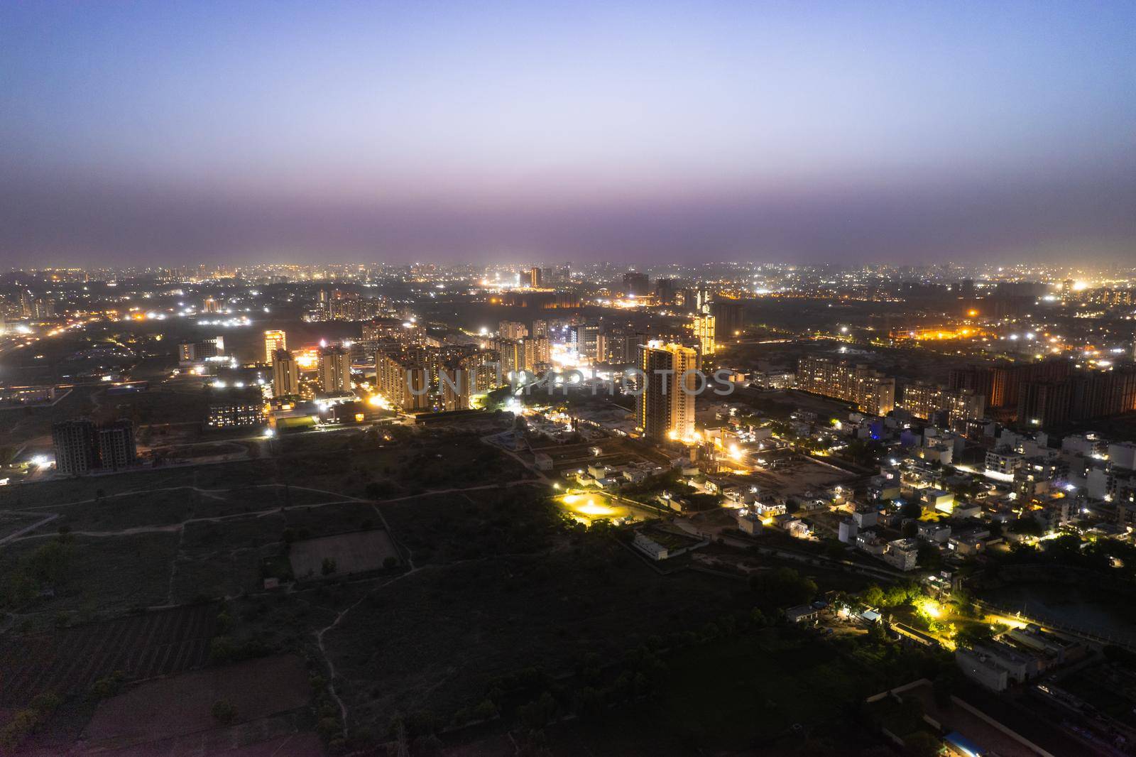 aerial drone dusk twilight shot showing orange lights of streets, homes and markets surrounding a skyscraper with the city scape stretching into the distance in gurgaon haryana delhi drone shot