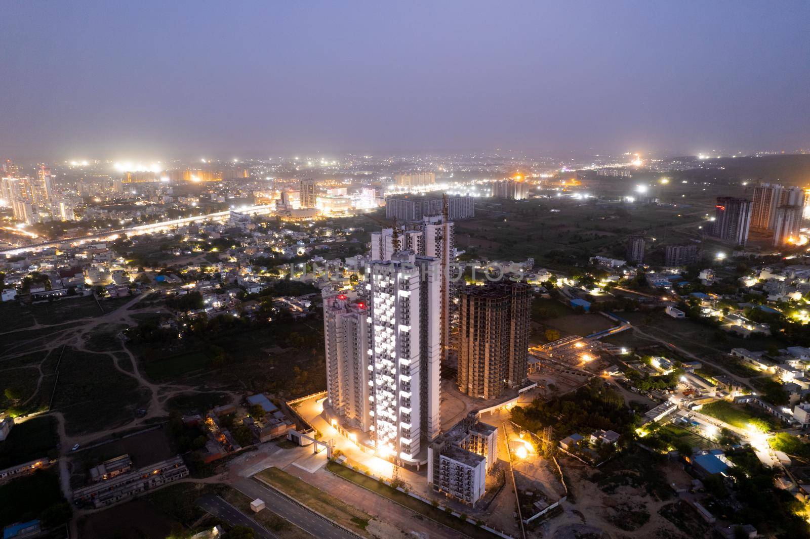 aerial drone dusk twilight shot showing orange lights of streets, homes and markets surrounding a skyscraper with the city scape stretching into the distance in gurgaon haryana delhi drone shot