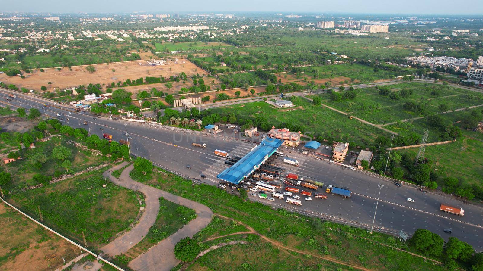 Aerial drone flying forward shot showing congestion traffic at toll booth on national highway in India with cars, trucks and vehicles waiting to cross the blue roofed structure with green trees all around by Shalinimathur