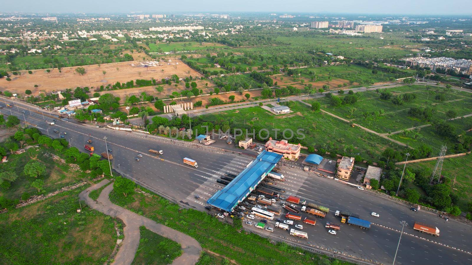 Aerial drone flying forward shot showing congestion traffic at toll booth on national highway in India with cars, trucks and vehicles waiting to cross the blue roofed structure with green trees all around by Shalinimathur