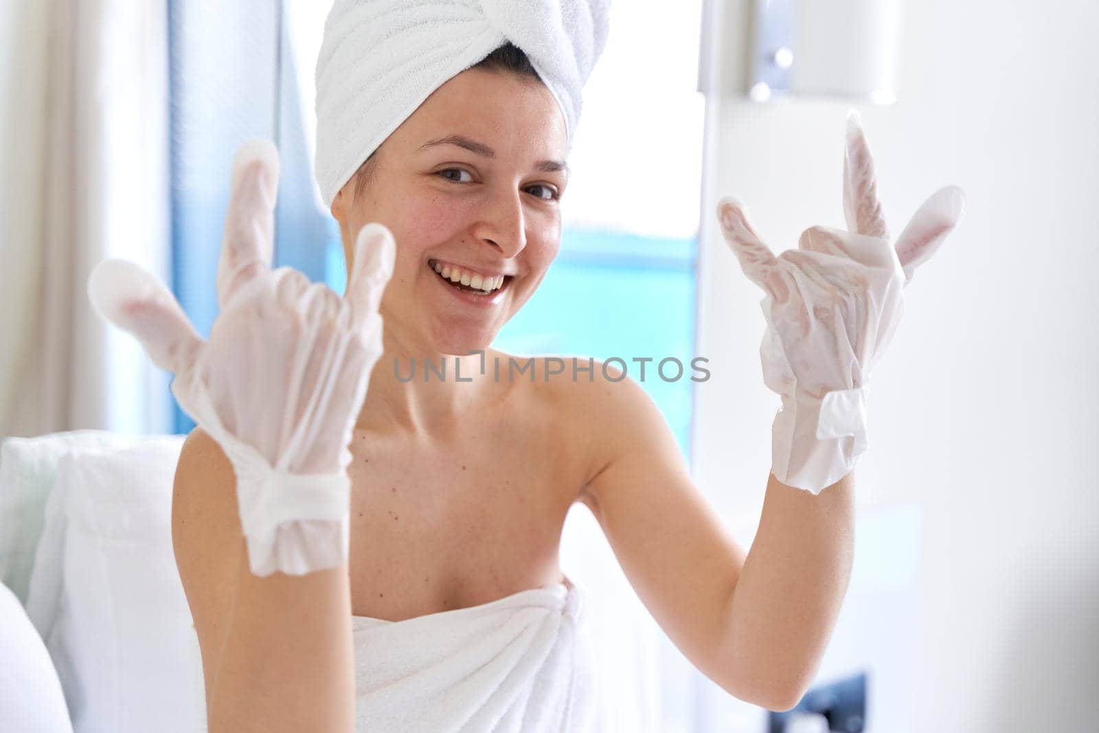 After a shower, a girl wrapped in a towel uses cosmetic gloves to moisturize the skin of her hands. Cosmetic trends for body care at home.