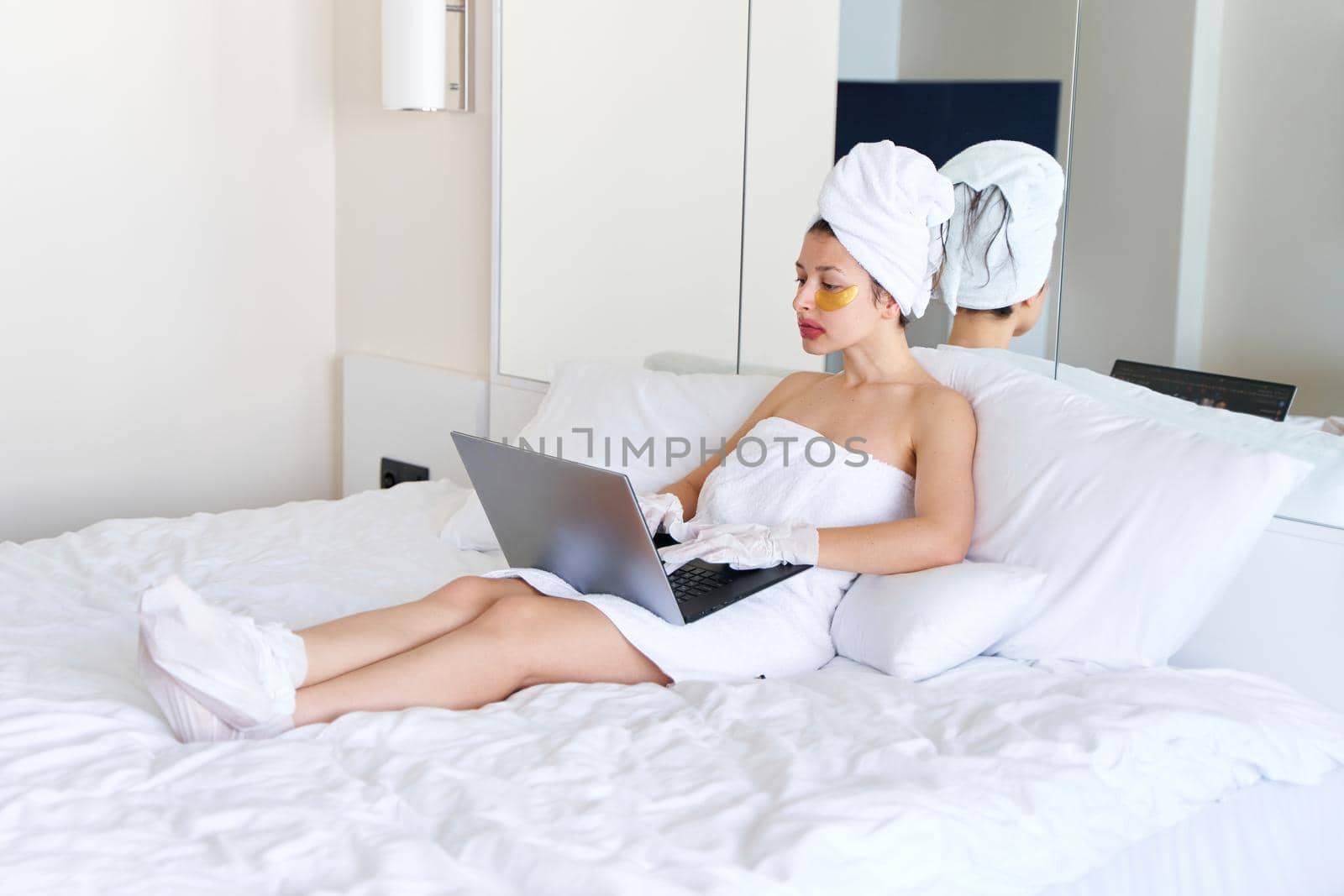 After a shower, a girl wrapped in a towel works on a laptop and uses cosmetic patches for the skin under the eyes, lips and gloves to moisturize her hands and feet. Cosmetic trends for body care at home.