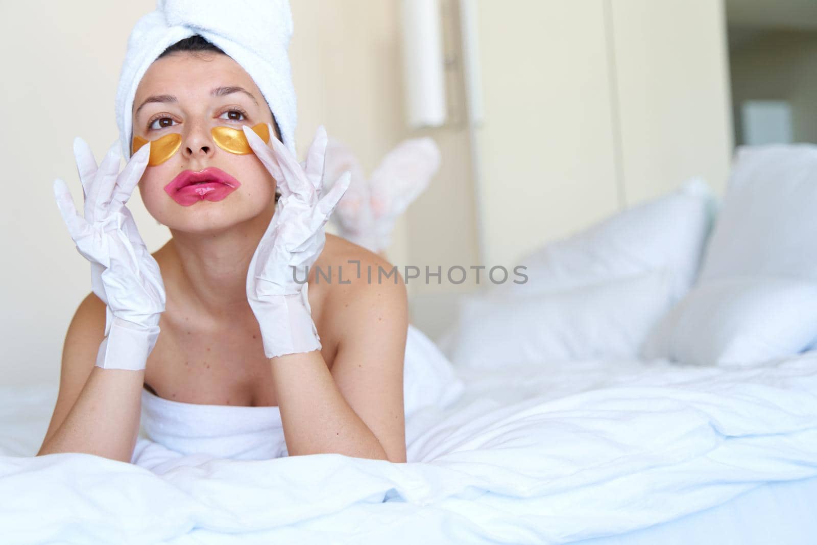 After a shower, a girl wrapped in a towel uses cosmetic patches for the skin under the eyes, lips and gloves to moisturize her hands and feet. Cosmetic trends for body care at home by Try_my_best