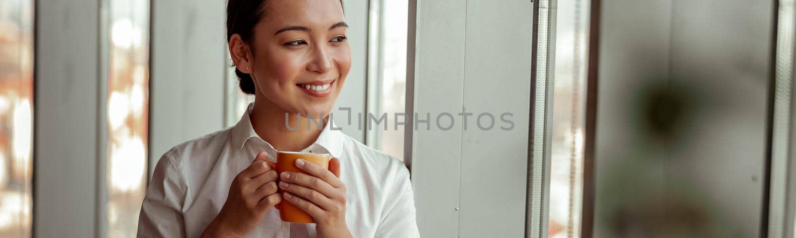Smiling asian business woman drinking coffee standing near window at office. High quality photo