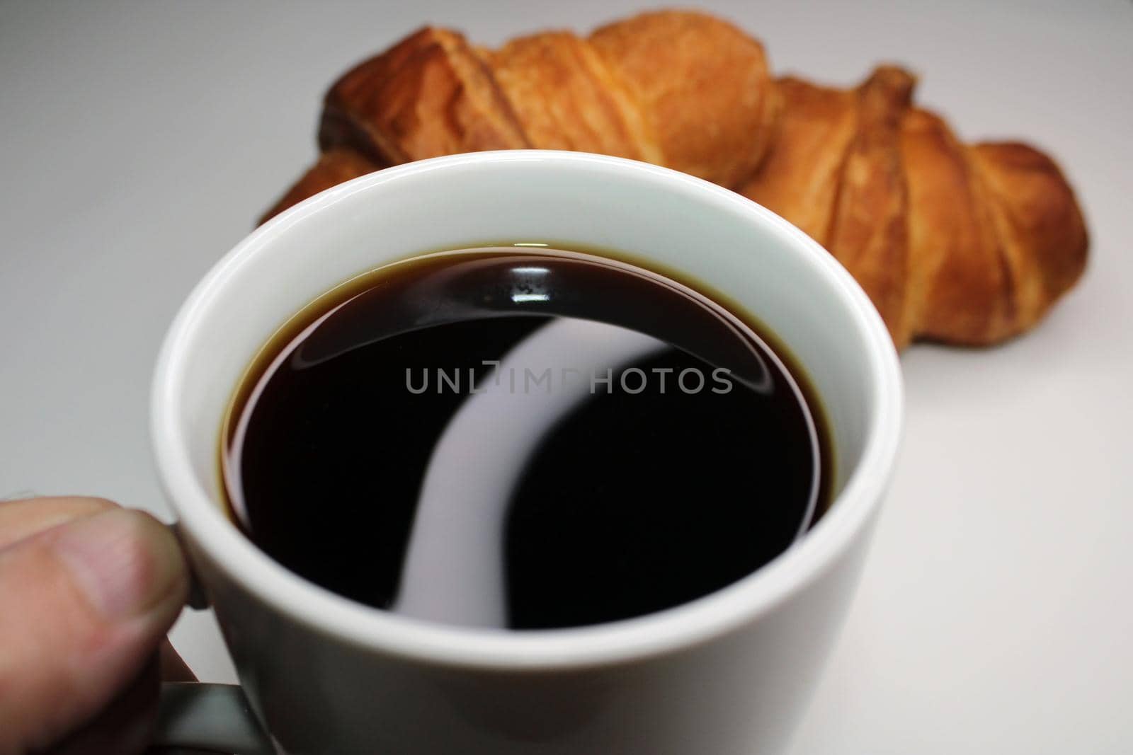 White cup with black coffee close-up on a light background. There are two croissants in the background. Cooking, home baking.