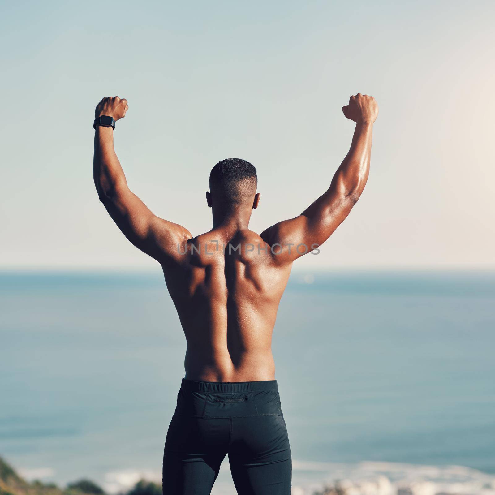 Hes the strongest one there is. Rearview shot of an unrecognizable young man standing with his hands raised while exercising outdoors. by YuriArcurs