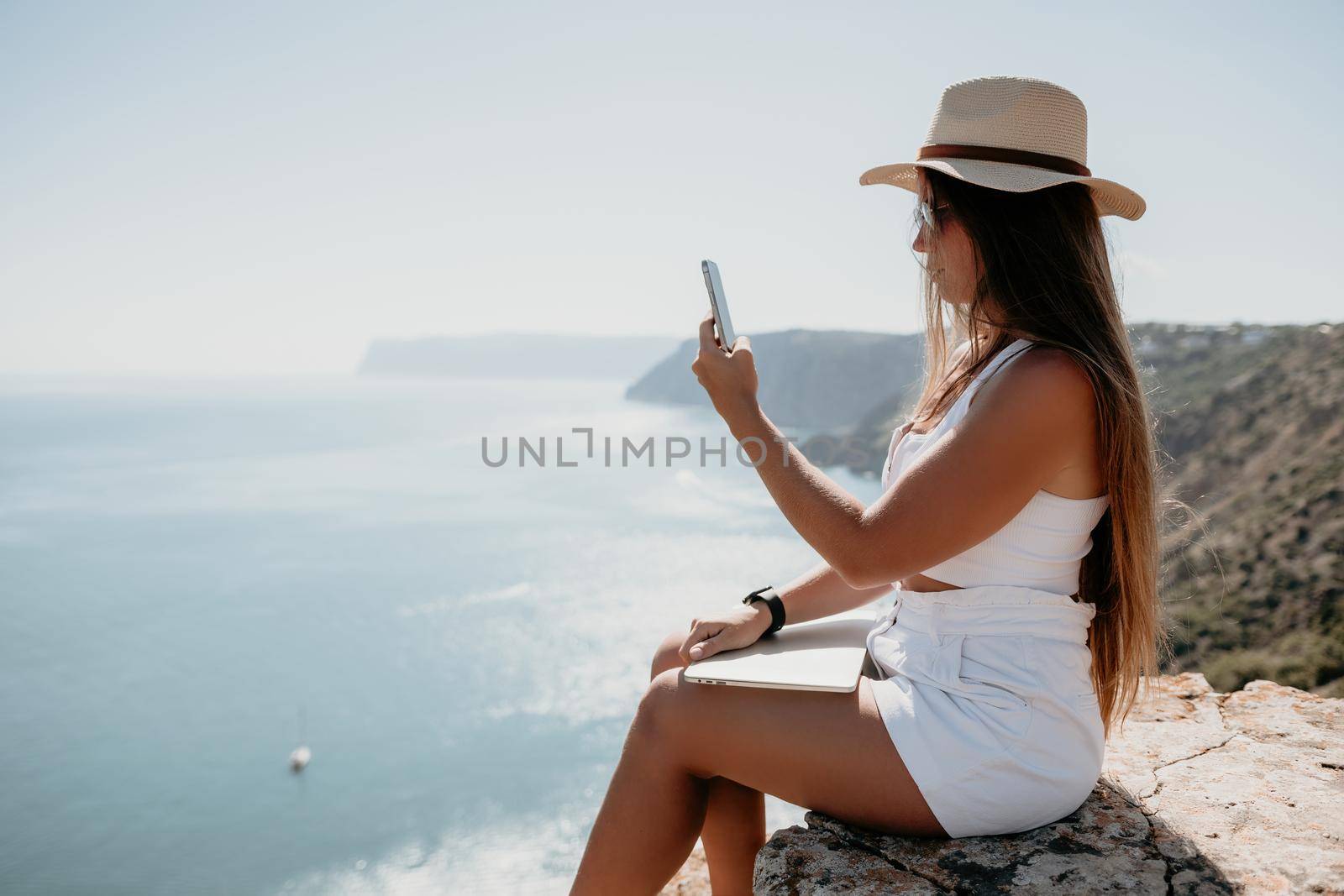 Successful business woman in yellow hat working on laptop by the sea. Pretty lady typing on computer at summer day outdoors. Freelance, travel and holidays concept.