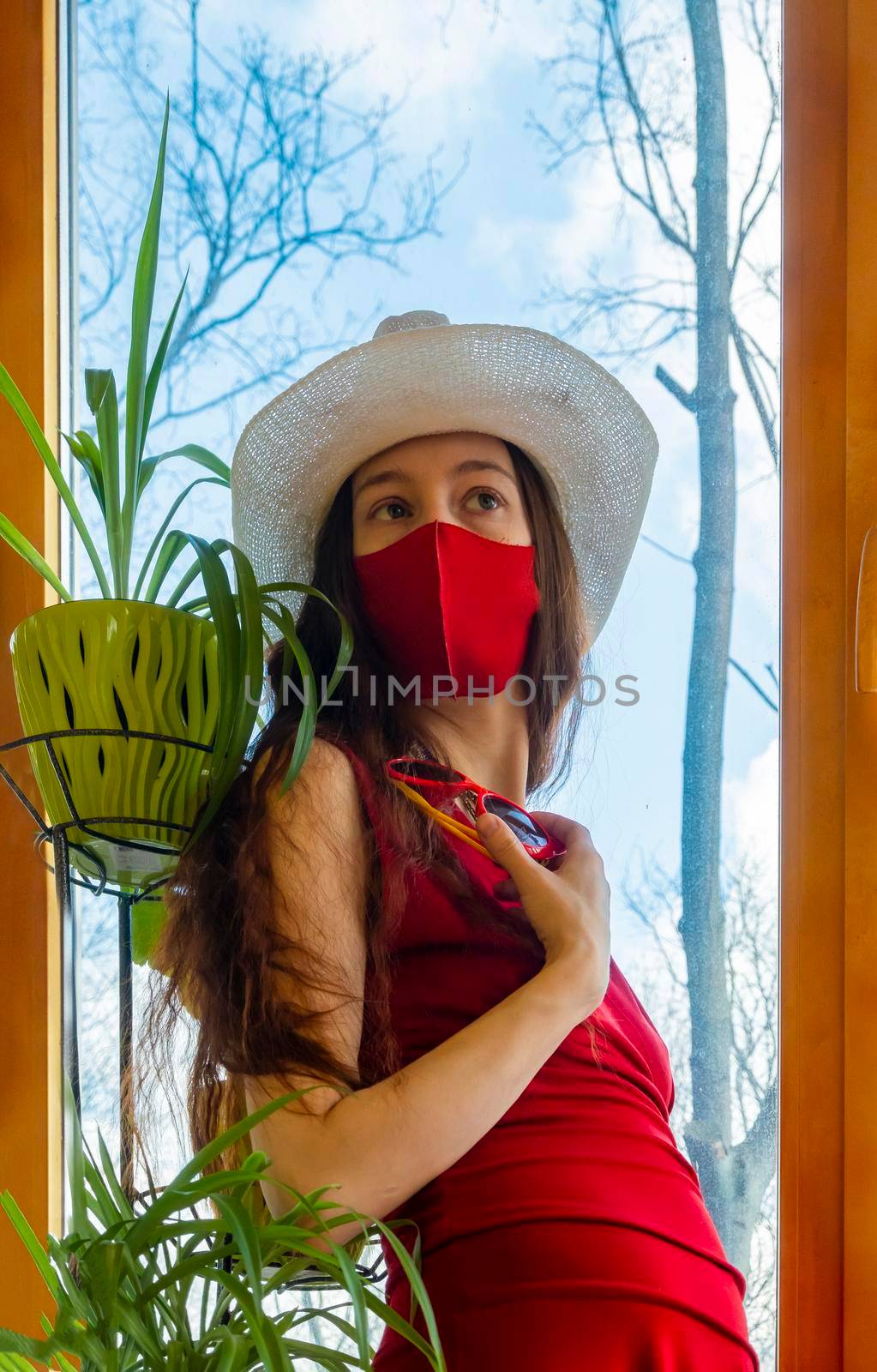A girl in a red dress, a white hat and a red medical mask is wearing funny sunglasses with plants against the sky in early spring