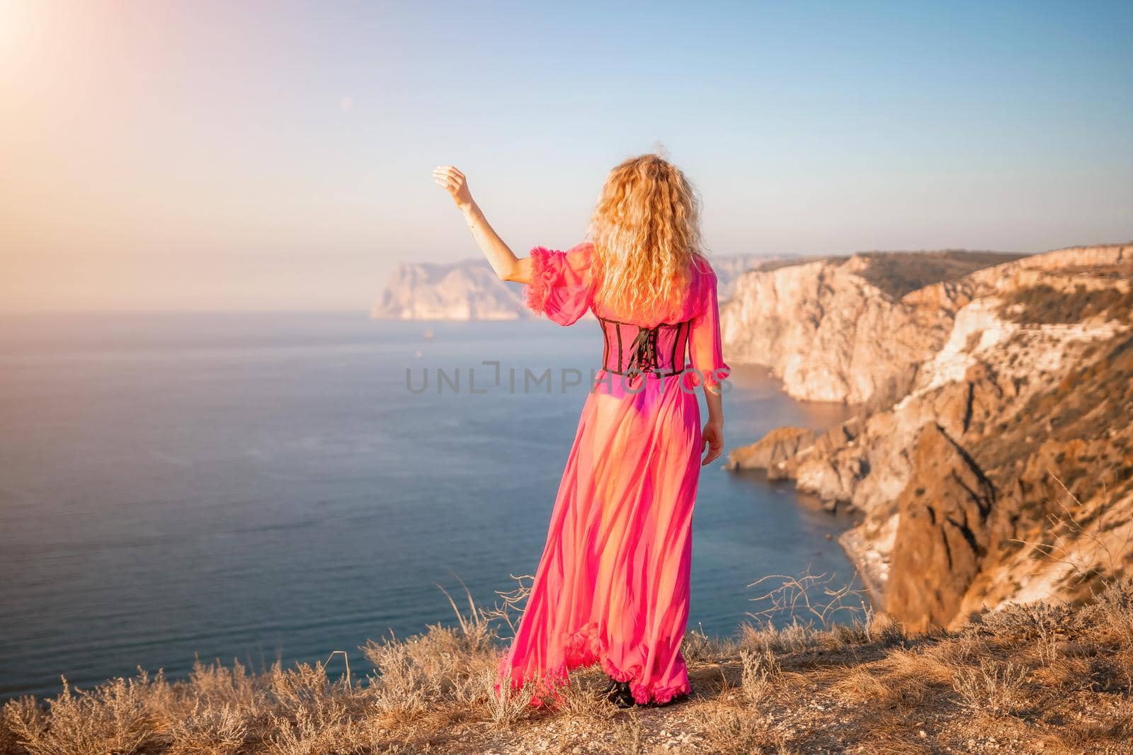 Curly redhead caucasian woman with freckles. Cute beautiful young blonde woman in a pink long dress posing on a volcanic rock high above the sea during sunset. Fashion travel conept by panophotograph