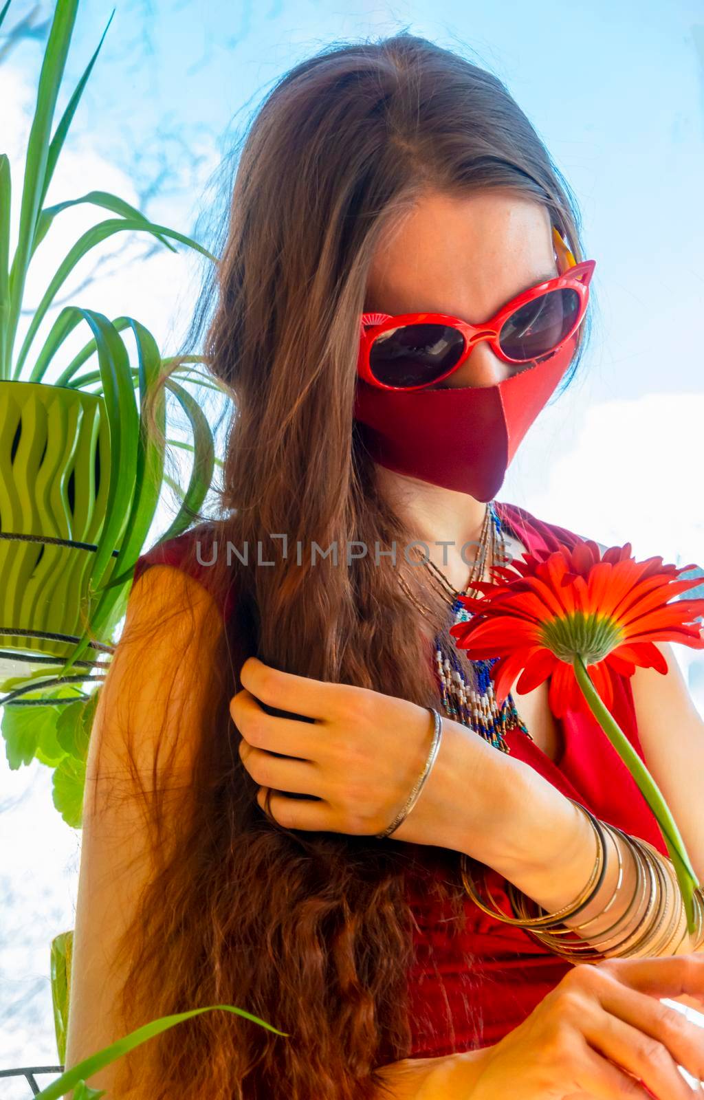 A girl in a red dress, red medical mask and funny sunglasses holds a red gerbera flower