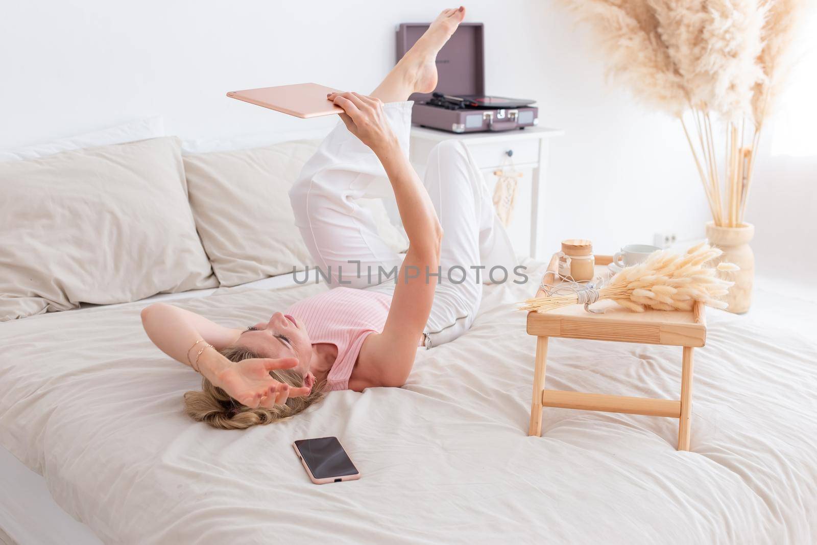 A slender beautiful woman, 30-40 years old, with blond hair, in a pink top and white pants, lies on the bed in the morning on her back, holds a digital tablet in her hands, a smartphone lies nearby