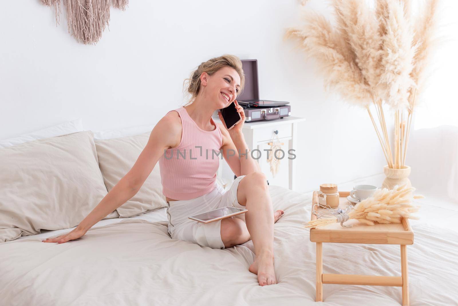 Happy woman with blonde hair, in a pink top and white pants, sitting on the bed in the morning, in a bright bedroom interior, calling on a smartphone