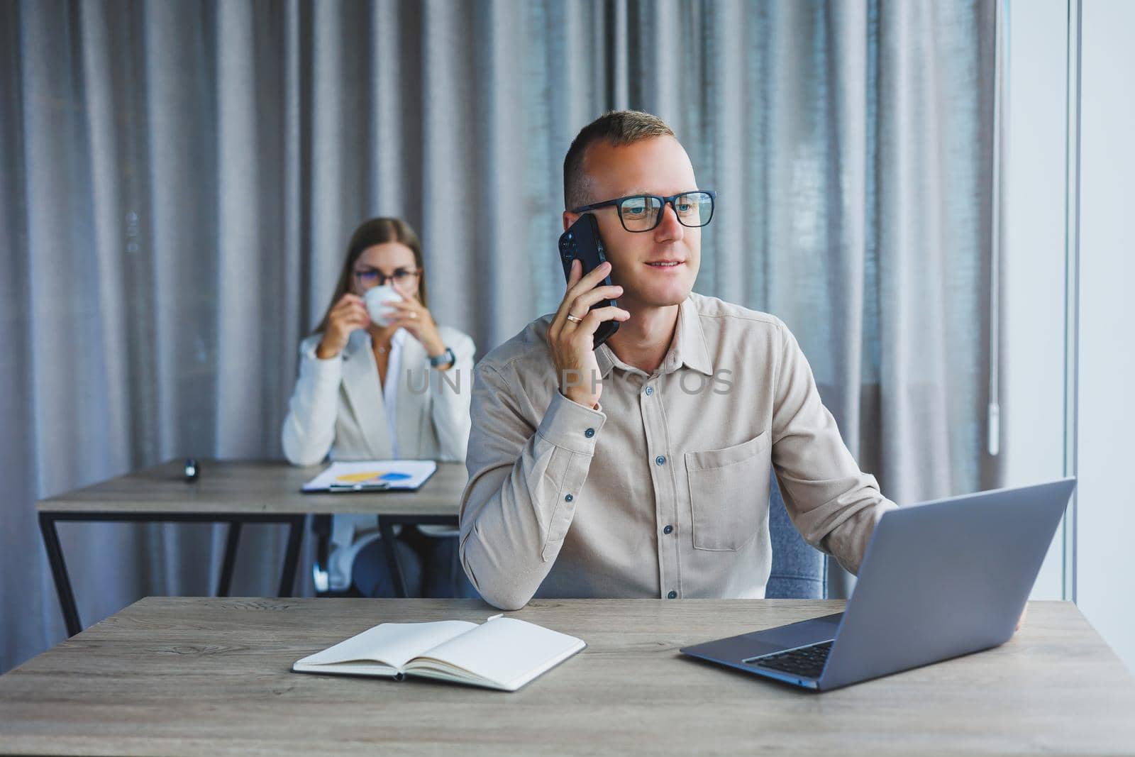 A male manager is talking on a mobile phone while sitting at a table with a computer and a notepad. Working atmosphere in an office with large windows