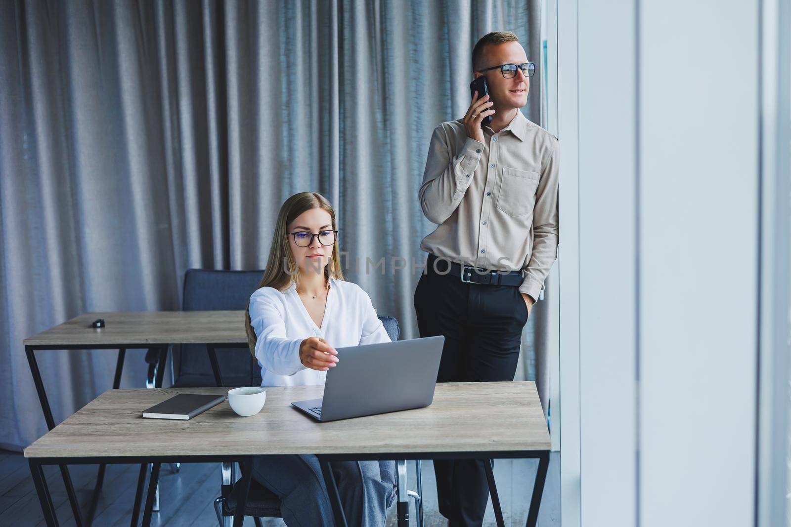 Confident male and female colleagues in elegant casual wear are discussing a project in the workplace, enjoying corporate friendship, happy partners are talking about office networks on a mobile phone