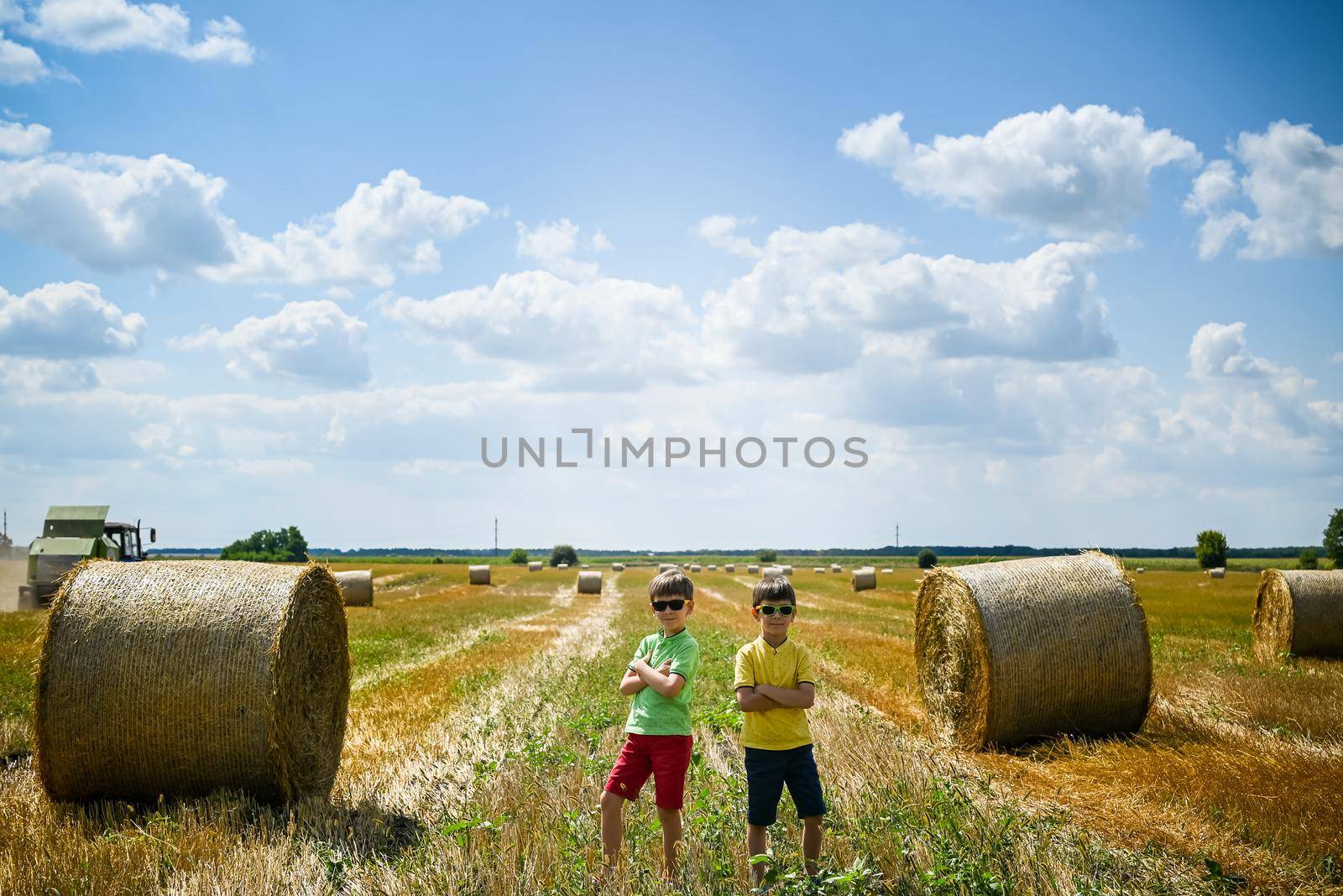 Two little boy stand among round haystack. Field with round bales after harvest under blue sky. Big round bales of straw, sheaves, haystacks.