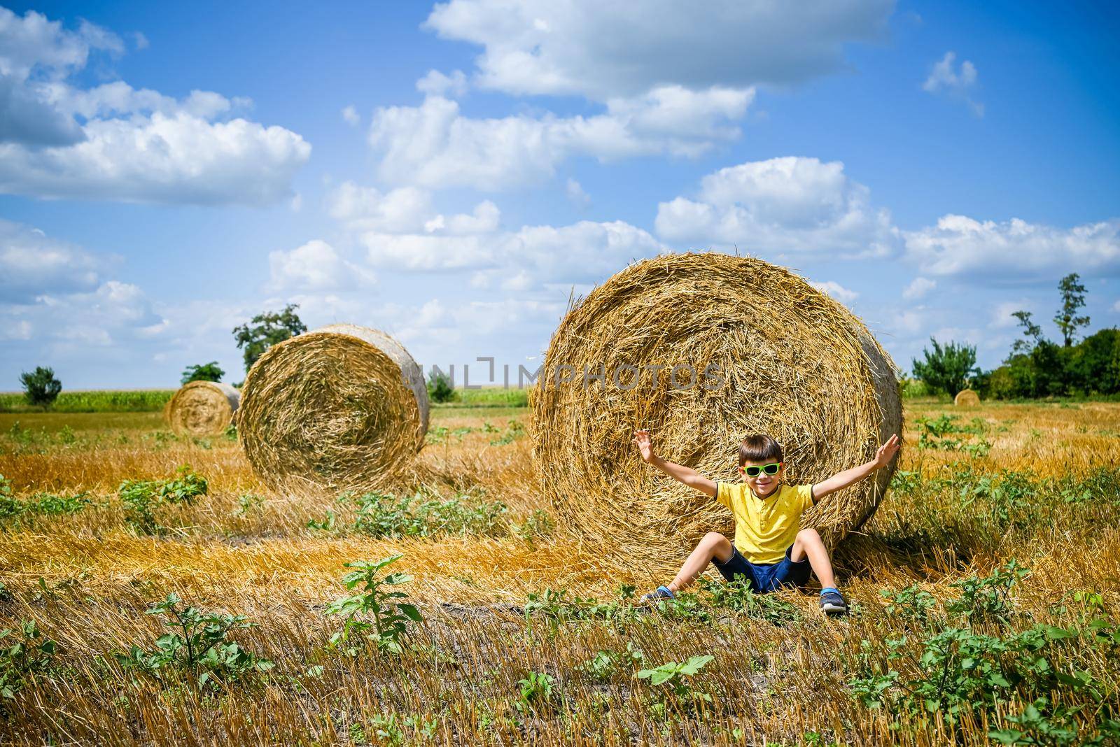 Little boy sits near a round haystack relaxed with hands up. Field with round bales after harvest under blue sky. Big round bales of straw, sheaves, haystacks.