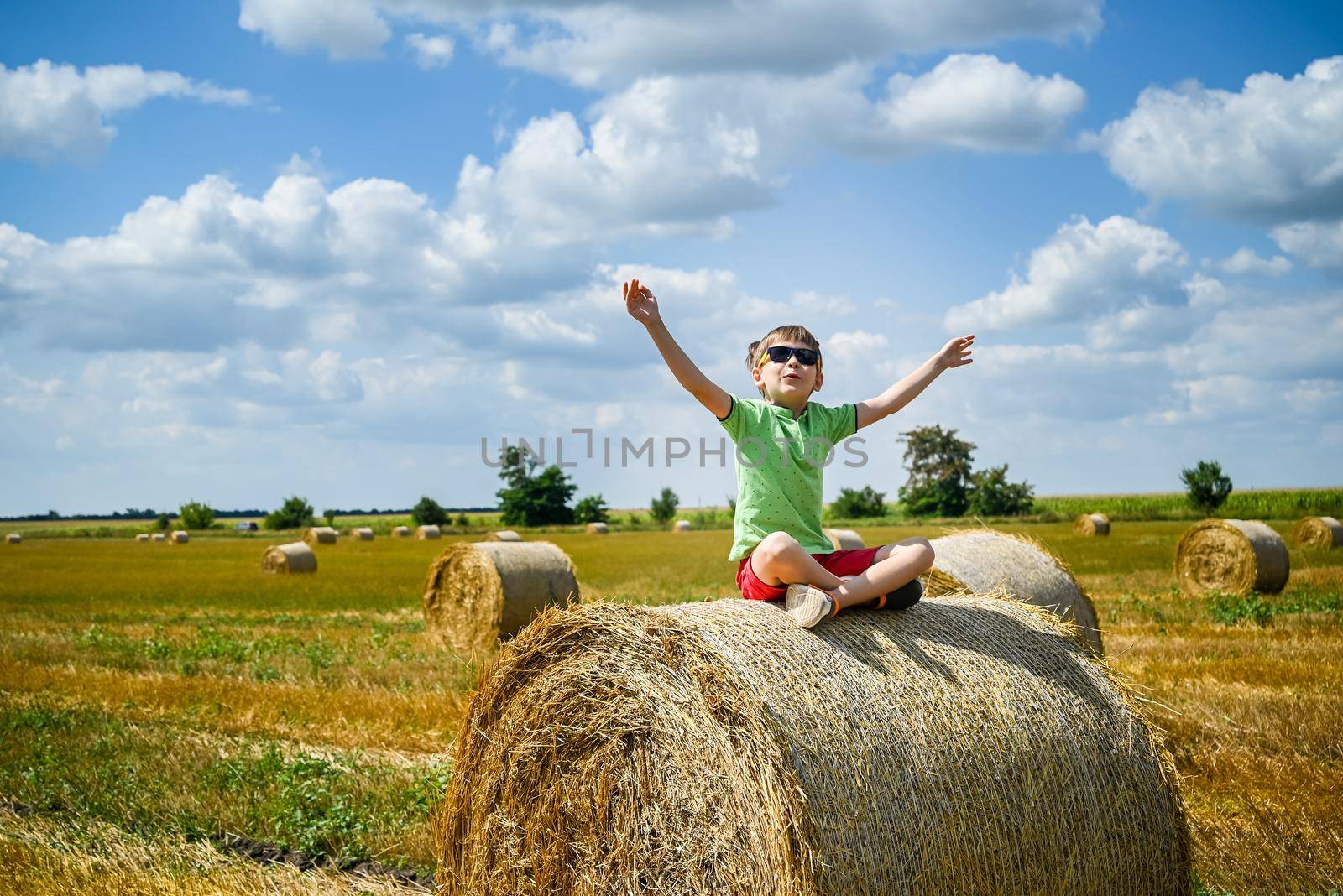 Little boy sits on a round haystack relaxed with hands up. Field with round bales after harvest under blue sky. Big round bales of straw, sheaves, haystacks.