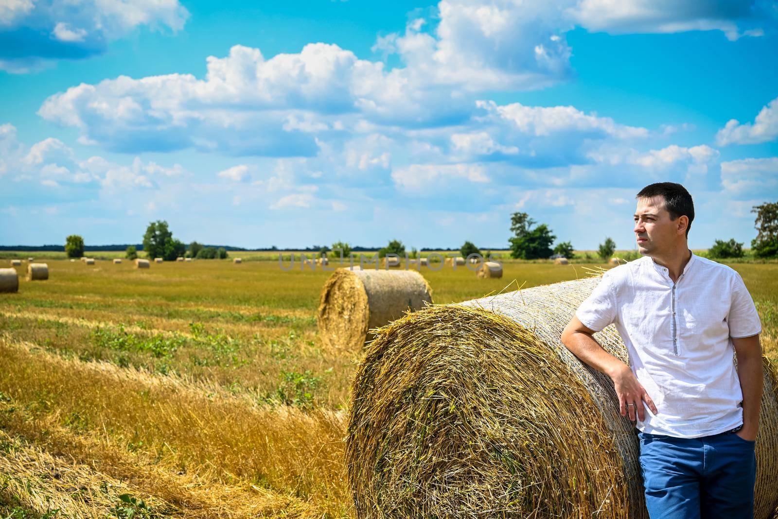 Happy farmer is standing beside bales of hay. He is examining straw after successful harvesting. Strong handsome man portrait on agricultural field.